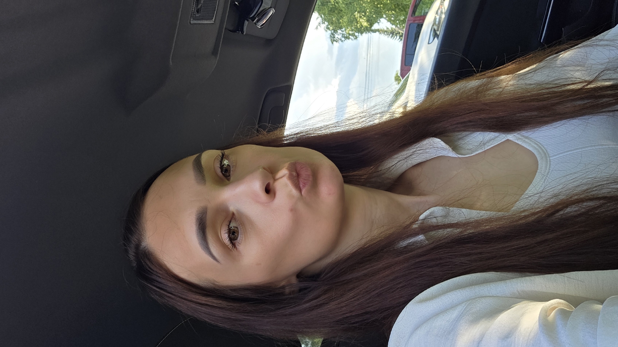 In the city of Bulgakovo, Russia, a young woman with long, dark brown hair is captured in a selfie taken inside a car. She is wearing a white collared shirt and a white ribbed tank top underneath. Her lips are pursed in a kiss pose, and she is looking directly at the camera. The interior of the car, including the dashboard and ceiling, is visible. Outside the car, through the window, hints of trees and a cloudy sky can be seen.