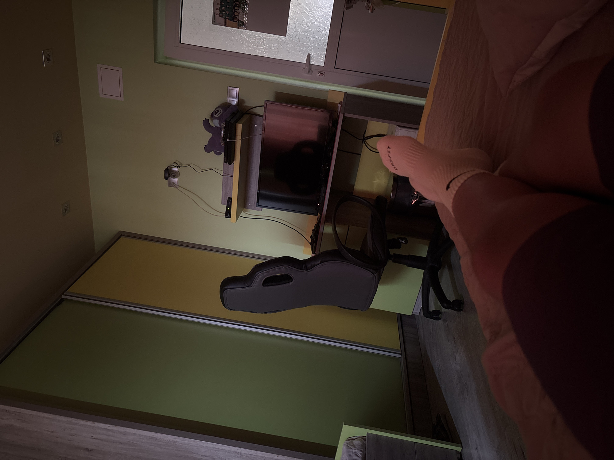 The image depicts an interior scene with a desk, computer setup, and part of a bed. A TV is situated on the desk, with a plush toy of Stitch to its left. Below the desk is a black computer chair. A portion of a bed with pink-toned bedding is visible on the right side of the image, with a person's legs and feet, wearing white socks, resting on the bed. The wall is painted a light yellow-green. There is a window or door visible at the top of the image. The presence of standard European power outlets suggests a location in Europe, and no location-specific items or landmarks are visible to confirm the city of Rakovski, Bulgaria. No specific actions or interactions are visible.