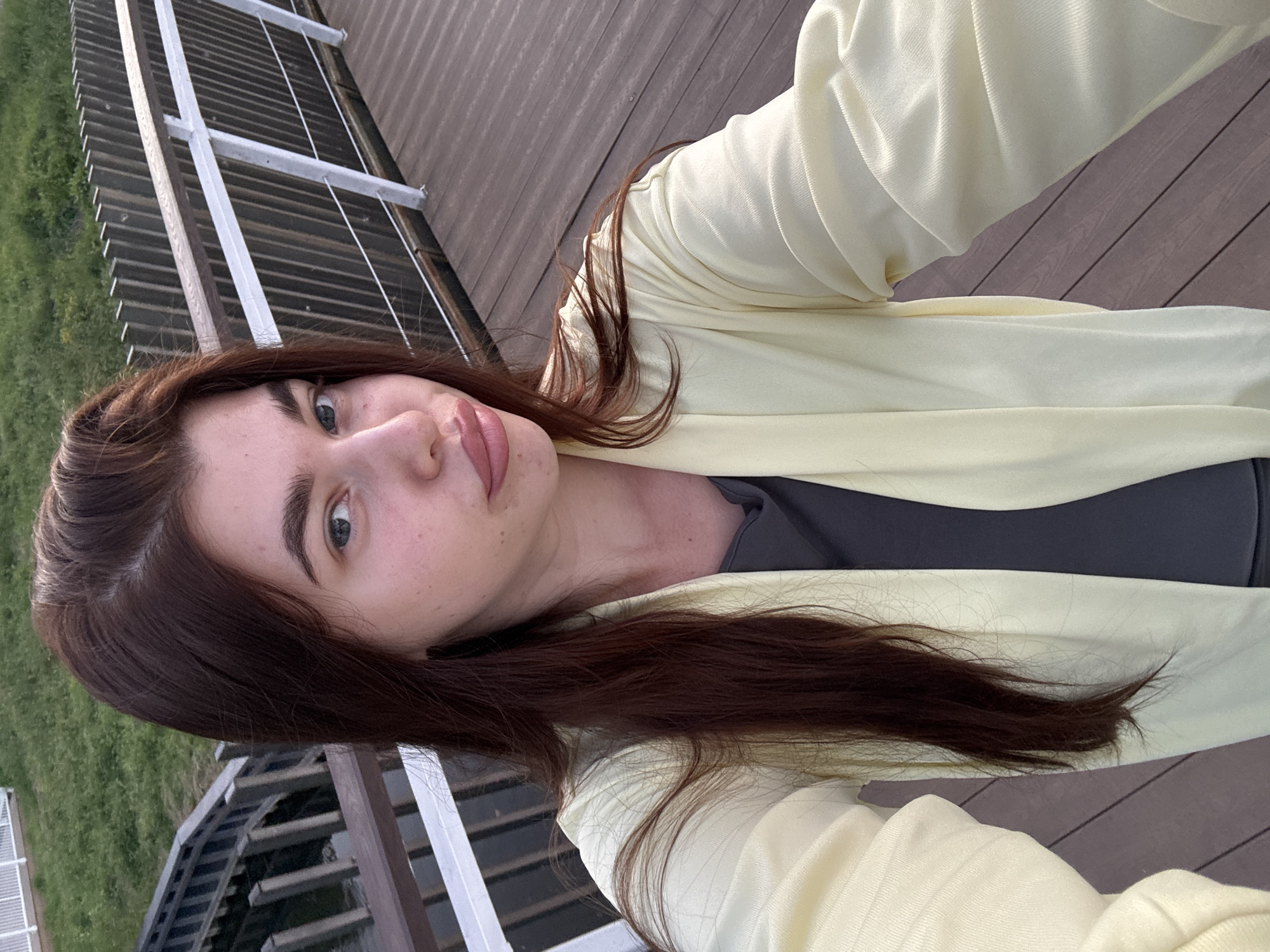 A young woman with long, dark brown hair and light blue eyes takes a selfie from a boardwalk. She is wearing a light yellow open cardigan over a dark grey t-shirt. The boardwalk is made of dark wood planks and has a white railing with metal supports. In the background, there is a grassy area and a wooden structure with steps leading down. The scene appears to be an outdoor recreational area, possibly a park or a waterfront. There is no identifiable information suggesting the location is Klin, Russia.