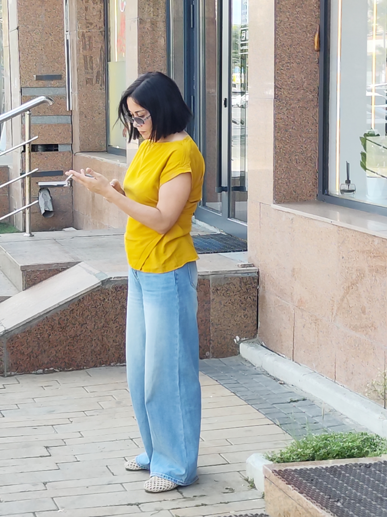 A woman with dark hair and sunglasses is standing on a paved walkway outside a building, holding a smartphone in her hands and looking down at it. She is wearing a yellow short-sleeved top and light blue wide-leg jeans. She has on light-colored woven flats. The building behind her has a textured brown facade and large windows. A metal railing is visible to her left, and stairs ascend to the left of the railing. The ground is paved with interlocking grey and beige stones. To the right of the woman, a small patch of green plants grows near a low wall. The scene appears to be in an urban setting, possibly Astana, Kazakhstan, given the context.