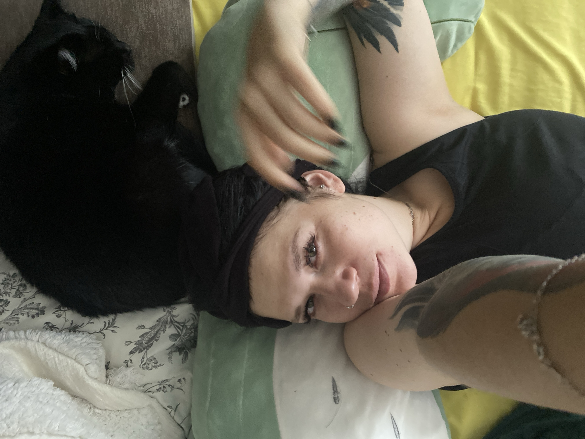 A selfie shows a woman with a black headband lying on her side. She has a nose ring and visible freckles. Her left arm is raised, with her hand blurred as if in motion. A black cat is visible in the background, sleeping on a patterned pillow. The woman is wearing a black tank top, and the scene appears to be indoors, possibly on a bed with yellow and green bedding. The image does not provide any information about the city of Chaikovsky, Russia.