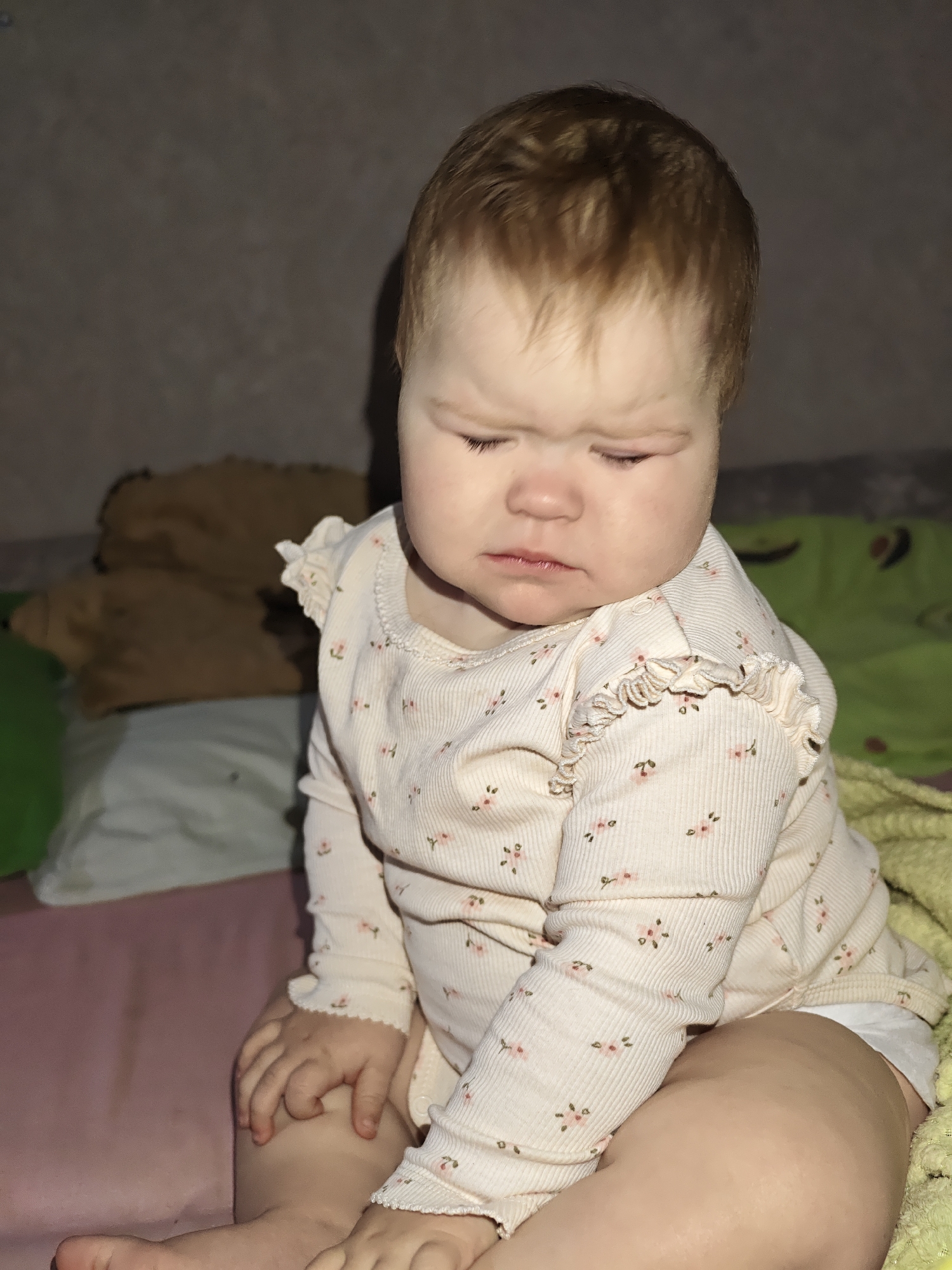 A baby with red hair is sitting indoors on a bed. The baby is wearing a long-sleeved, patterned shirt. The bed has a pink sheet and a green blanket. A brown stuffed animal is visible in the background. The specific location in Chaikovsky, Russia, cannot be determined from the image.