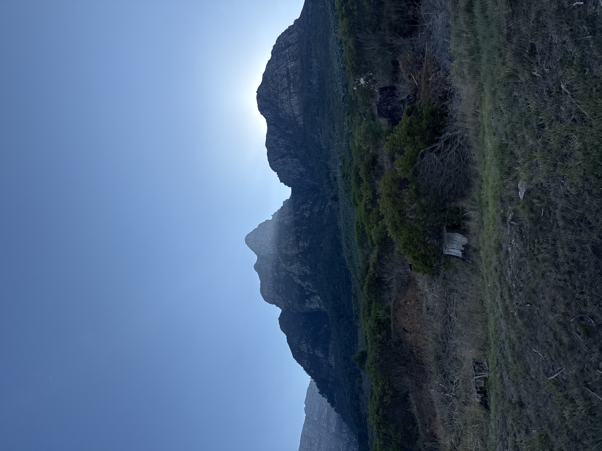 A landscape photograph shows a view in Cape Town, South Africa. A mountain range is illuminated from behind by the sun, which is obscured by the peak, creating a bright corona. The mountainside is covered with sparse green vegetation and dry grasses. The sky is clear and blue.