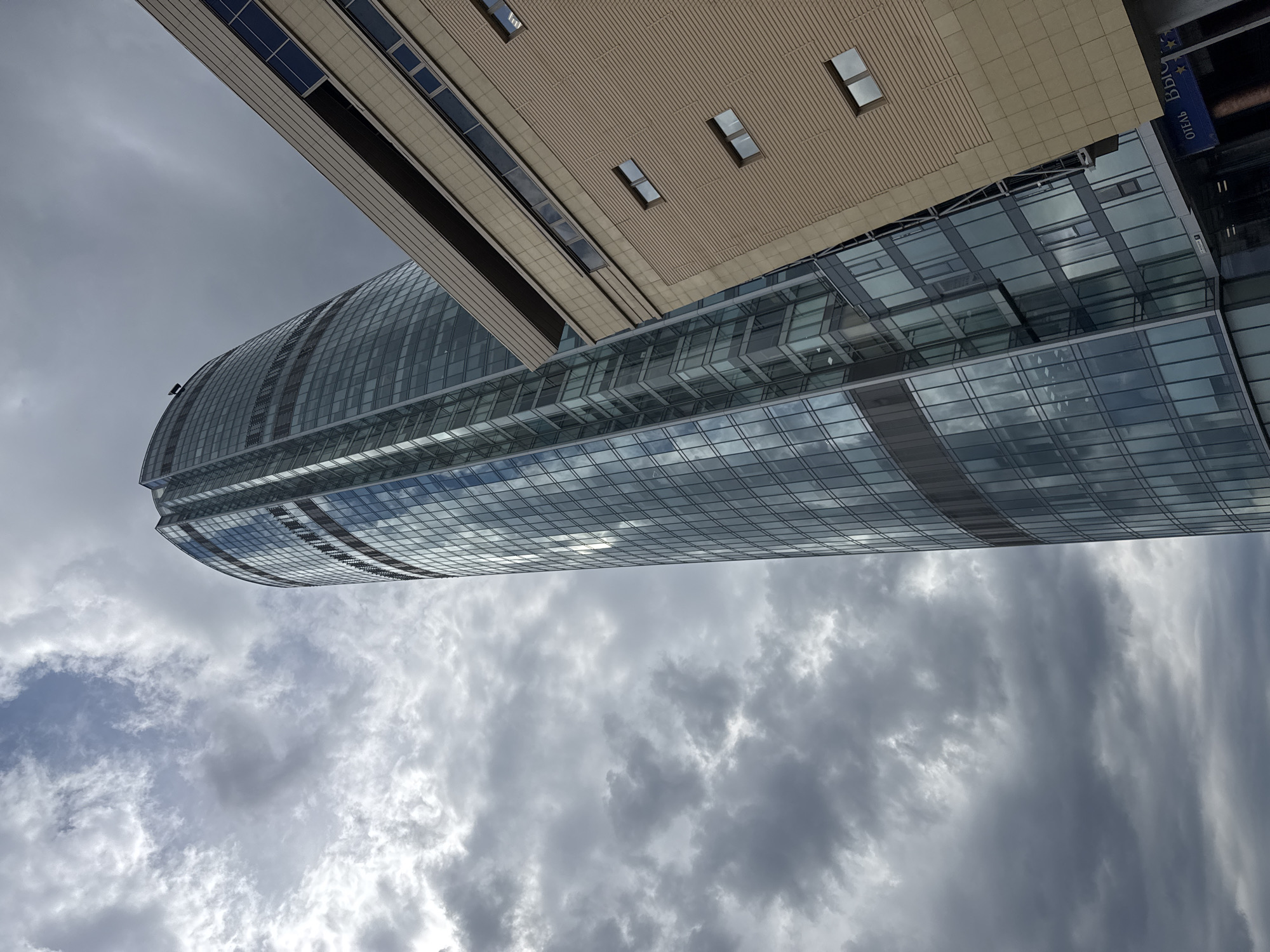 A low-angle, full shot captures the Vyssotsky skyscraper in Yekaterinburg, Russia, under a cloudy sky. The skyscraper, a tall, curved glass building, dominates the left side of the frame. To its right, a portion of a beige, horizontally paneled building is visible, with several rectangular windows. The sky is filled with dramatic, dark gray clouds, creating a moody atmosphere. A small sign with blue lettering and stars is visible at the base of the beige building. No people are present in the image.