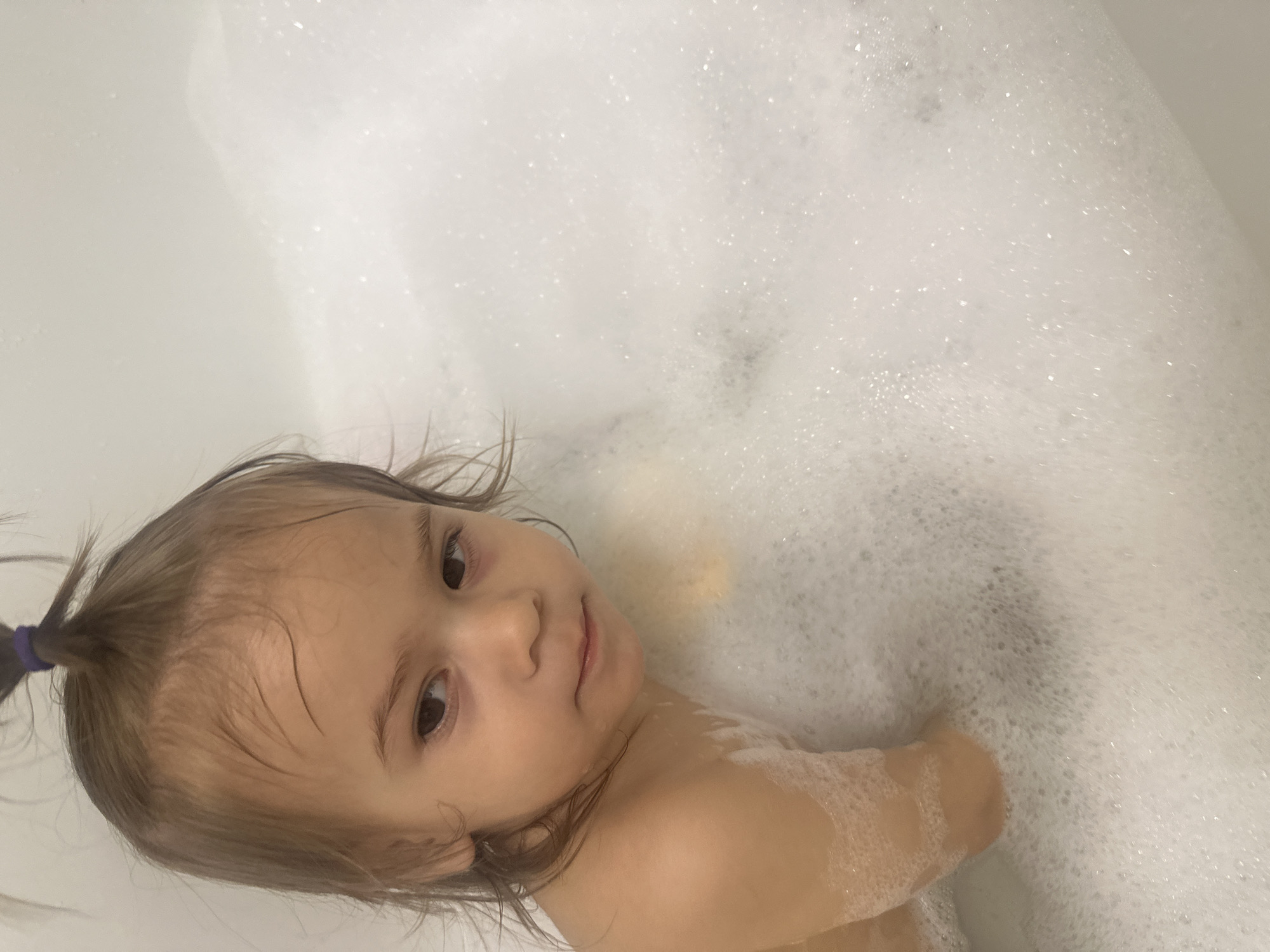 A young child with light brown hair, partially gathered in a ponytail with a dark blue band, is partially submerged in a bathtub filled with white bubbles. The child is looking towards the camera with a slight smile. The setting appears to be a private bathroom. The location is not discernible from the visual information provided.