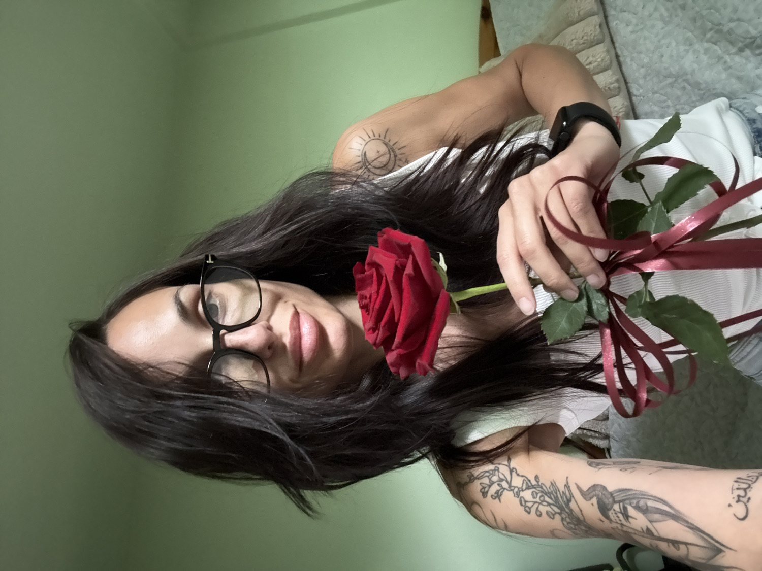 The image features a woman posing indoors, holding a single red rose. She is shown from the chest up, with her head tilted, looking directly at the camera with a gentle, slight smile. Her long, dark hair flows around her shoulders. She wears black-rimmed glasses and a white ribbed tank top.
In her right hand, she holds a vibrant red rose, whose stem is wrapped with a dark red ribbon tied into a bow. A black digital watch is visible on her left wrist. Her arms and shoulder display several tattoos. On her left shoulder, there is a circular design, possibly depicting a sun and moon. Her right forearm is heavily tattooed with a botanical motif of leaves and branches, a detailed bird-like creature, and some script in Arabic characters.
The background behind her head is a plain light green wall, while a soft, textured white or light grey material, possibly a cushion or blanket, is visible behind her torso. The lighting appears soft and even, suggesting an indoor setting. The scene takes place in Pleven, Bulgaria, and the woman's expression conveys a calm and perhaps appreciative emotion.