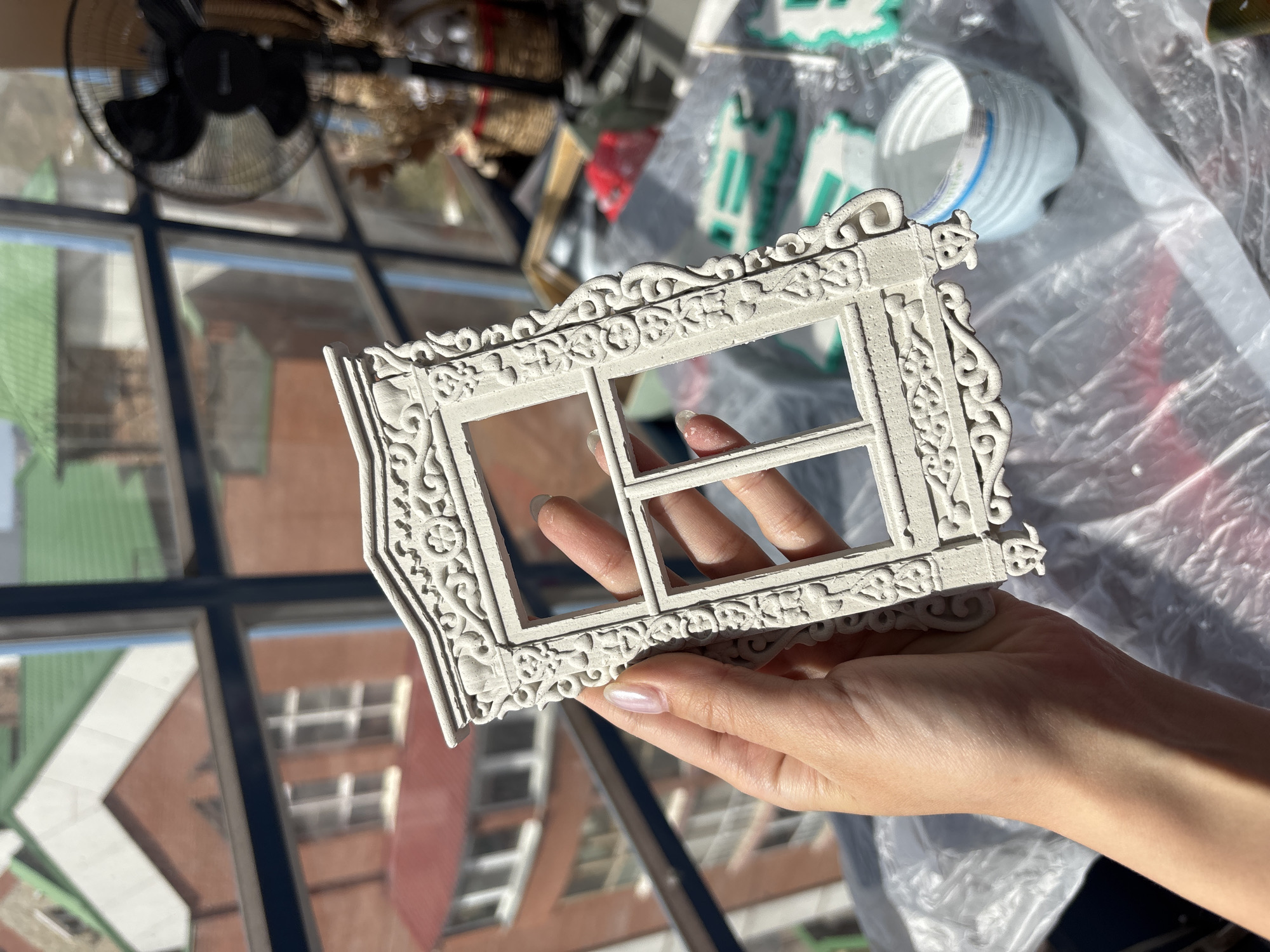 A hand holds a small, white, ornate window frame up to a large multi-paned window. The location appears to be in Krasnoyarsk, Russia, as a building with a green roof and reddish-brown brick or siding is visible through the window. There are other objects visible on a surface behind the hand including a plastic container, a fan, and other decorative items. The surface is covered with a piece of plastic.