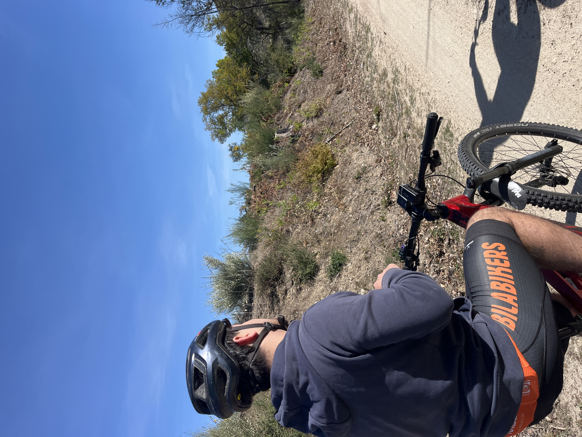A man wearing a helmet, grey hoodie, and cycling shorts with "BILABIKERS" printed on them is riding a red mountain bike on a dirt trail. The sky is blue and clear. The surrounding area has low-lying vegetation and trees. The location appears to be in a natural area near Vila Real, Portugal.