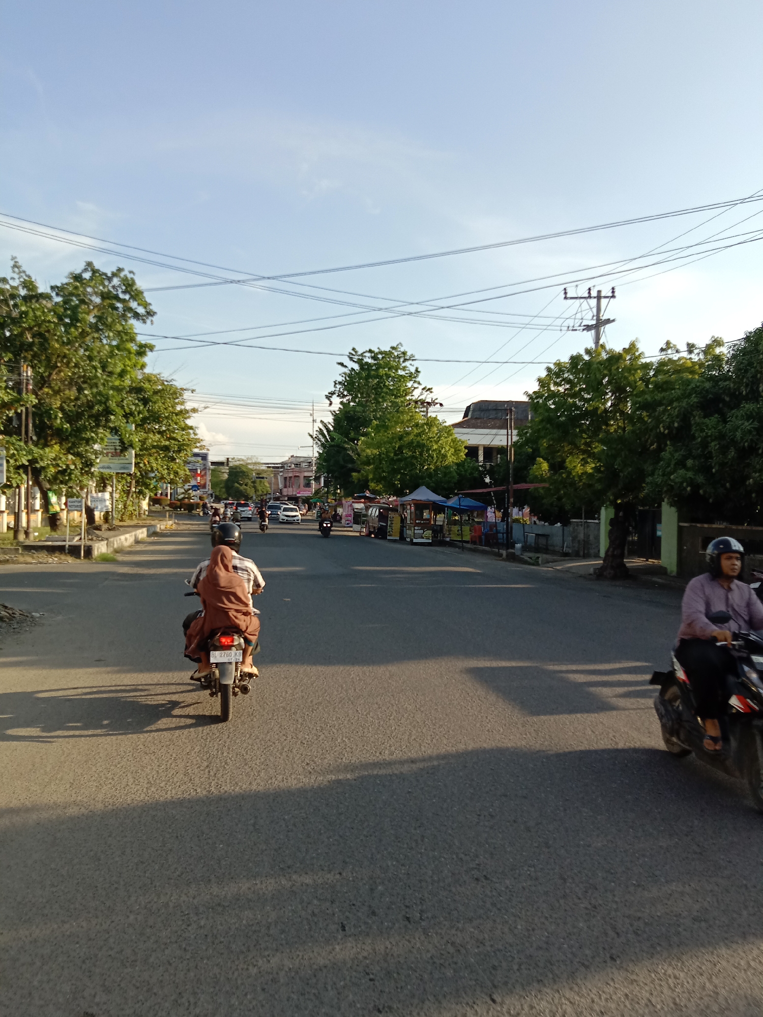 The image captures a street scene, likely in an urban or semi-urban area, consistent with Lhokseumawe, Indonesia. The perspective is from the road, looking down a straight street.
**Subjects:** The foreground features two motorcyclists. One, seen from behind, wears a brown hijab and a plaid shirt, with a passenger also in brown. Their motorcycle has a license plate "BL 2760 KB" and "07 24" visible below it. To the right, another person wearing a dark helmet and a purple long-sleeved shirt rides a black motorcycle. Further down the road, additional motorcycles and a few cars are visible, indicating traffic. The street is lined on both sides with lush green trees. On the right, several street vendor stalls with blue and white awnings are set up. Utility poles with a dense network of overhead power lines crisscross the sky. Buildings of various sizes and styles are visible behind the trees and stalls.
**Activity or event:** The main activity is people commuting or traveling on motorcycles along the street. The presence of vendor stalls suggests daily commerce or a market area.
**Notable details:** The time of day appears to be either early morning or late afternoon, indicated by the long shadows cast across the asphalt road by the motorcycles and trees, and the warm, low-angle sunlight. The sky is largely clear and light blue. The scene depicts a typical street environment in a Southeast Asian town, characterized by motorcycle dominance and roadside vendors.
**Visible text:** The most legible text is the license plate "BL 2760 KB" with "07 24" on the motorcycle in the left foreground. On a white sign on the left side of the road, the word "KANTOR" (Indonesian for "office") is partially visible. Other signs are too blurry to read.