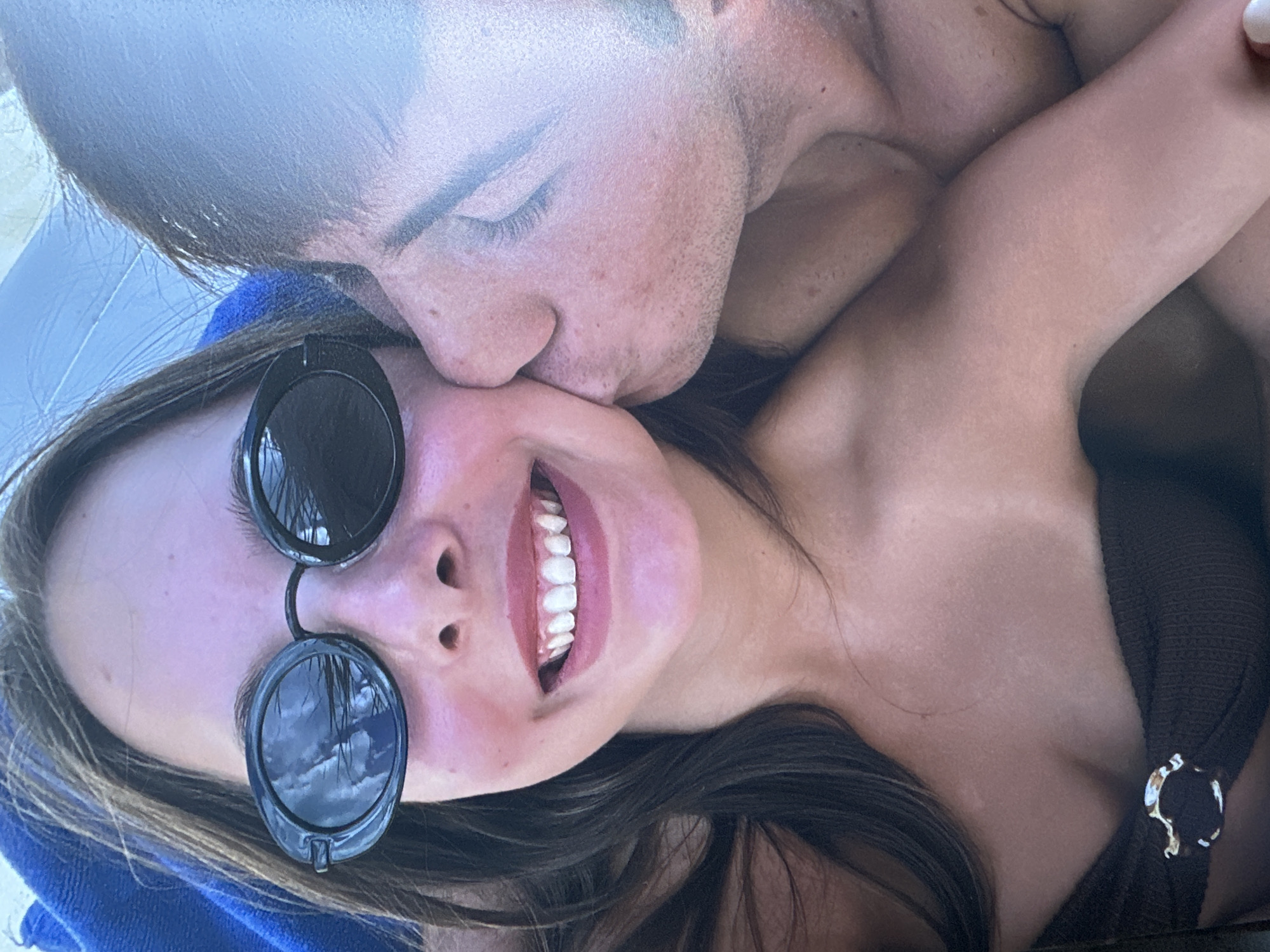A close-up, eye-level, selfie-style shot captures a young woman with shoulder-length brown hair and black, oval-shaped sunglasses. She is smiling, revealing white teeth, and has a light dusting of blush on her cheeks. A man's head is close to her face, with his lips touching her cheek. The man has stubble on his face and closed eyes. The background shows a blue towel and what appears to be an umbrella. The lower portion of the image shows the woman's chest, where she is wearing a brown bikini top with a decorative buckle. The overall setting appears to be outdoors, possibly at a beach or poolside. There is no indication of the city of Sofia, Bulgaria, in the image.