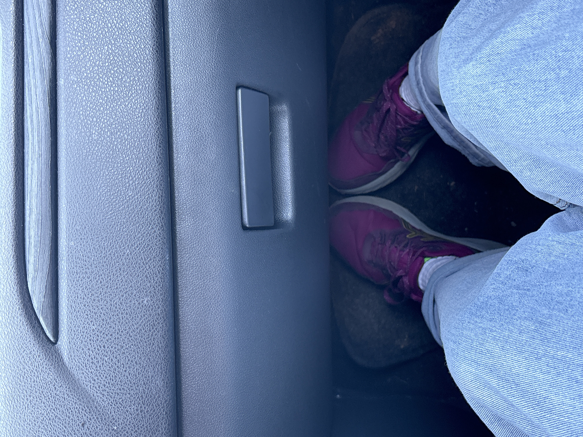 The image depicts a person sitting in the passenger seat of a car. The interior of the car is visible, with a focus on the dashboard and the person's legs. The person is wearing blue jeans and purple sneakers. The dashboard is a textured gray plastic with a silver handle. The floor mat is dark gray. The location is unknown.