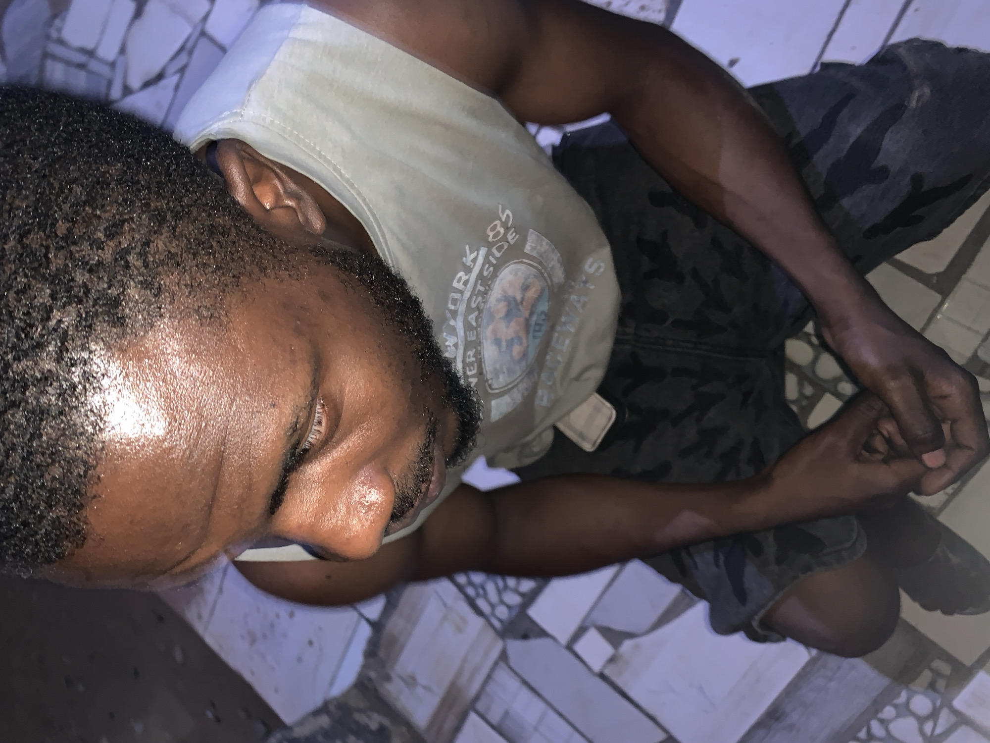 A man is lying on his side on a patterned tile floor. He is wearing a light-colored t-shirt with a circular graphic and camouflage pants. His dark hair is short, and he has a trimmed beard. He is clenching his hands. The location is in the city of New Stadium, Nigeria.