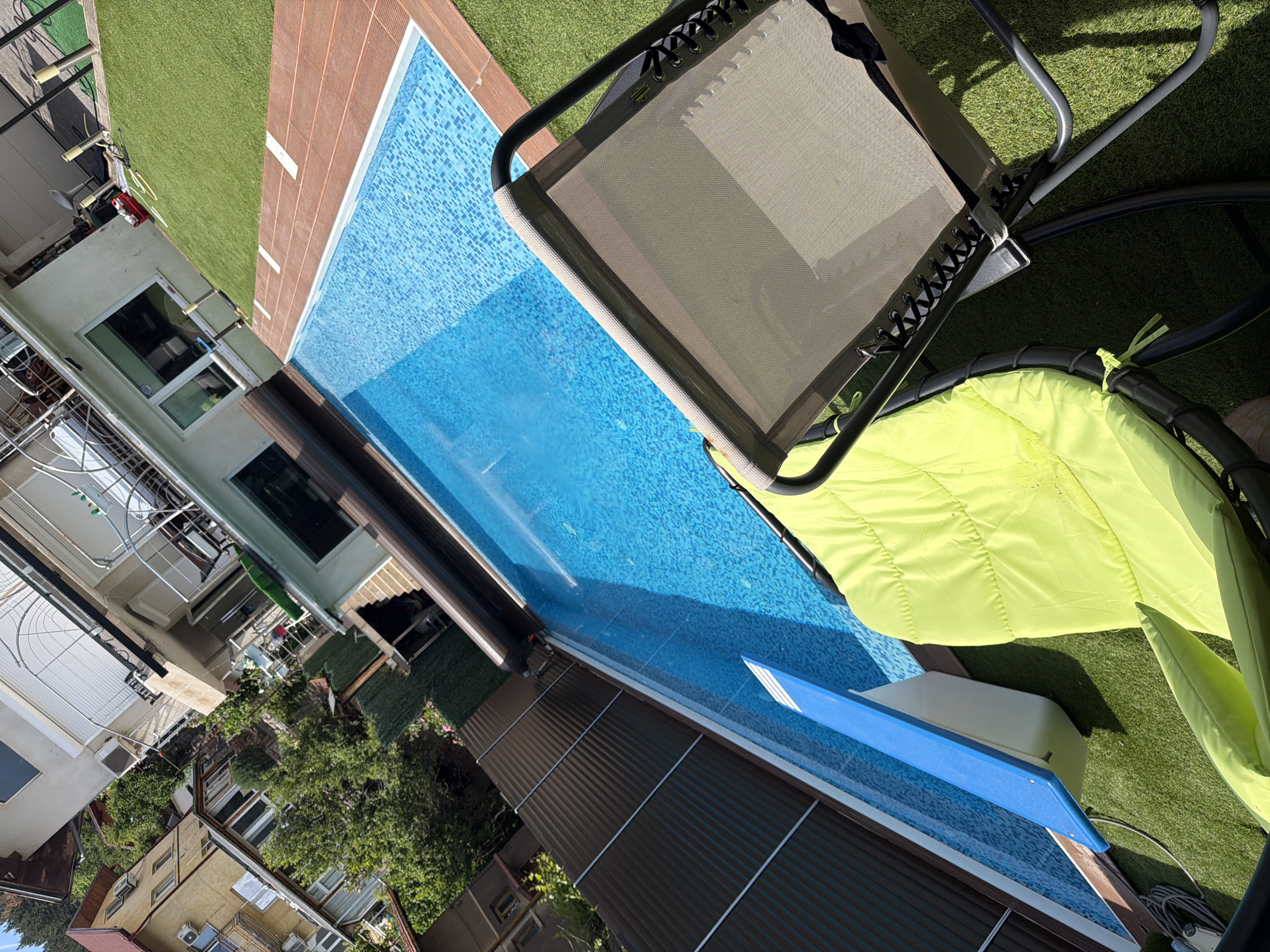 The image depicts a residential backyard with a swimming pool located in Sofia, Bulgaria. The pool is rectangular with a blue tiled bottom and is partially covered by a brown retractable cover. The surrounding area is covered with artificial turf. In the foreground, there are two chairs: one is a bright green cushioned lounge chair, and the other is a grey metal lounge chair with a mesh seat. Behind the pool, a two-story house with a beige facade and white trim is visible, featuring balconies and windows. To the left of the house, other residential buildings and dense green trees are present. The scene appears to be a private residence designed for leisure and recreation.