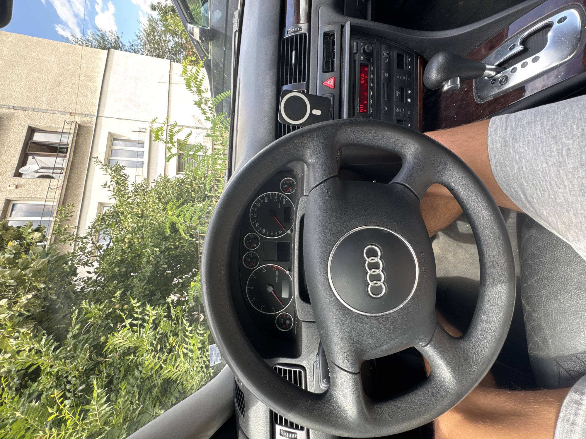 A first-person perspective shot from the driver's seat of an Audi vehicle shows the steering wheel, dashboard, and part of the passenger seat. The car is parked outdoors, with lush green foliage and a light-colored, multi-story building visible through the windshield. The building appears to be an apartment complex, with several windows and a satellite dish on one of the upper floors. The sky is blue with scattered white clouds.
The dashboard features a multi-function steering wheel with the Audi logo prominently displayed in the center. To the right of the steering wheel is the car's radio, which is currently tuned to FM 1 at 107.8 MHz. A hazard light button is visible above the radio. There is a magnetic phone mount attached to one of the air vents. The gear shift, for an automatic transmission, is visible to the right of the steering wheel. The interior of the car shows signs of wear and dust. The driver's legs, clad in grey shorts, are visible in the lower portion of the frame, suggesting the photo was taken while seated in the driver's seat.
The presence of the building's architecture, the style of the apartment building, and the overall urban setting strongly suggest the location is within Sofia, Bulgaria.
