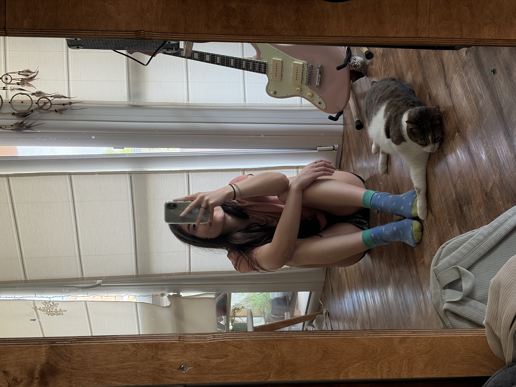 A young woman is taking a mirror selfie in an indoor setting. She is sitting on a wooden floor with her legs crossed, wearing a pink t-shirt, black shorts, and blue and green socks. She is holding a smartphone in her right hand, capturing her reflection. A tabby and white cat is lying on the floor to her right. Behind the woman, a pale pink electric guitar stands upright on a stand. The background features large windows with blinds, a dreamcatcher hanging from the ceiling, and a hint of houseplants reflected in the mirror. The overall environment appears to be a living space.