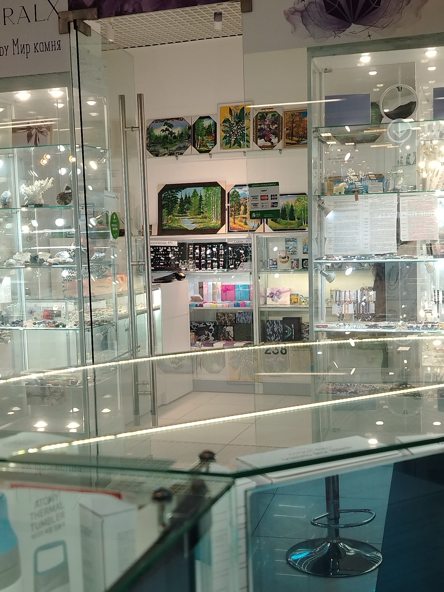 A brightly lit retail space, likely a jewelry or gift shop, is visible in the city of Izhevsk, Russia. The shop features glass display cases filled with merchandise, including stones, jewelry, and other decorative items. Several framed art pieces depicting landscapes and foliage hang on the wall. The shop's name, "RALX," is partially visible at the top of the frame, alongside the Russian phrase "Ду Мир камня," suggesting a focus on the world of stones. A barcode or code labeled "238" is noticeable. A bar stool stands in the foreground, reflecting light. A box labeled "ATONIY THERMAL TUMBLER" is partially visible in the immediate foreground. The overall environment is clean and well-lit, suggesting a retail setting.