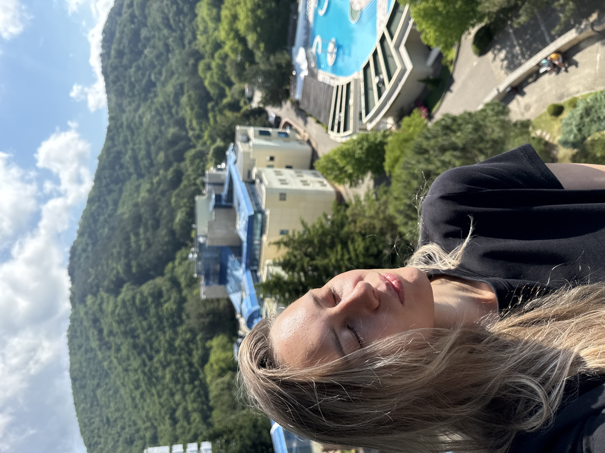 An outdoor, medium shot captures a young woman from the chest up, her head tilted back with eyes closed, basking in the sun. She has long, blonde hair and wears a black t-shirt. The background reveals a lush green mountainside under a bright blue sky with scattered clouds. Below the mountain, a resort complex with modern buildings, including structures with blue roofs, is visible, along with a vibrant blue swimming pool. The location is identified as Nebug, Russia.