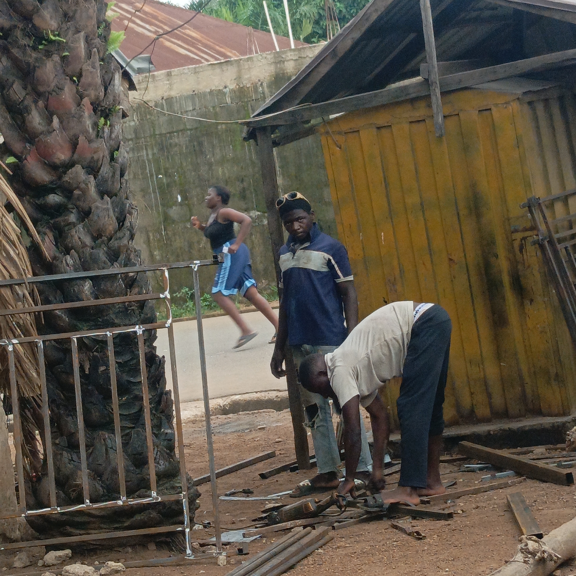 In Akwanga, Nigeria, the image captures a scene on a street where two men are engaged in metalwork. One man, wearing a light-colored shirt and dark pants, is bent over, operating a power tool on a piece of metal. He is barefoot. The other man, standing and observing, is wearing a blue shirt with a white stripe and light-colored pants, also with sandals. He has sunglasses perched on his head. The metal pieces are scattered on the ground around them, suggesting an active workshop. A partially constructed metal gate stands nearby. A woman is jogging in the background on the street, wearing a black tank top and a blue skirt. Behind them, a weathered yellow structure with a partially collapsed roof is visible, next to a weathered concrete wall. A palm tree trunk dominates the left foreground. The scene indicates a roadside metalworking operation in an urban environment.