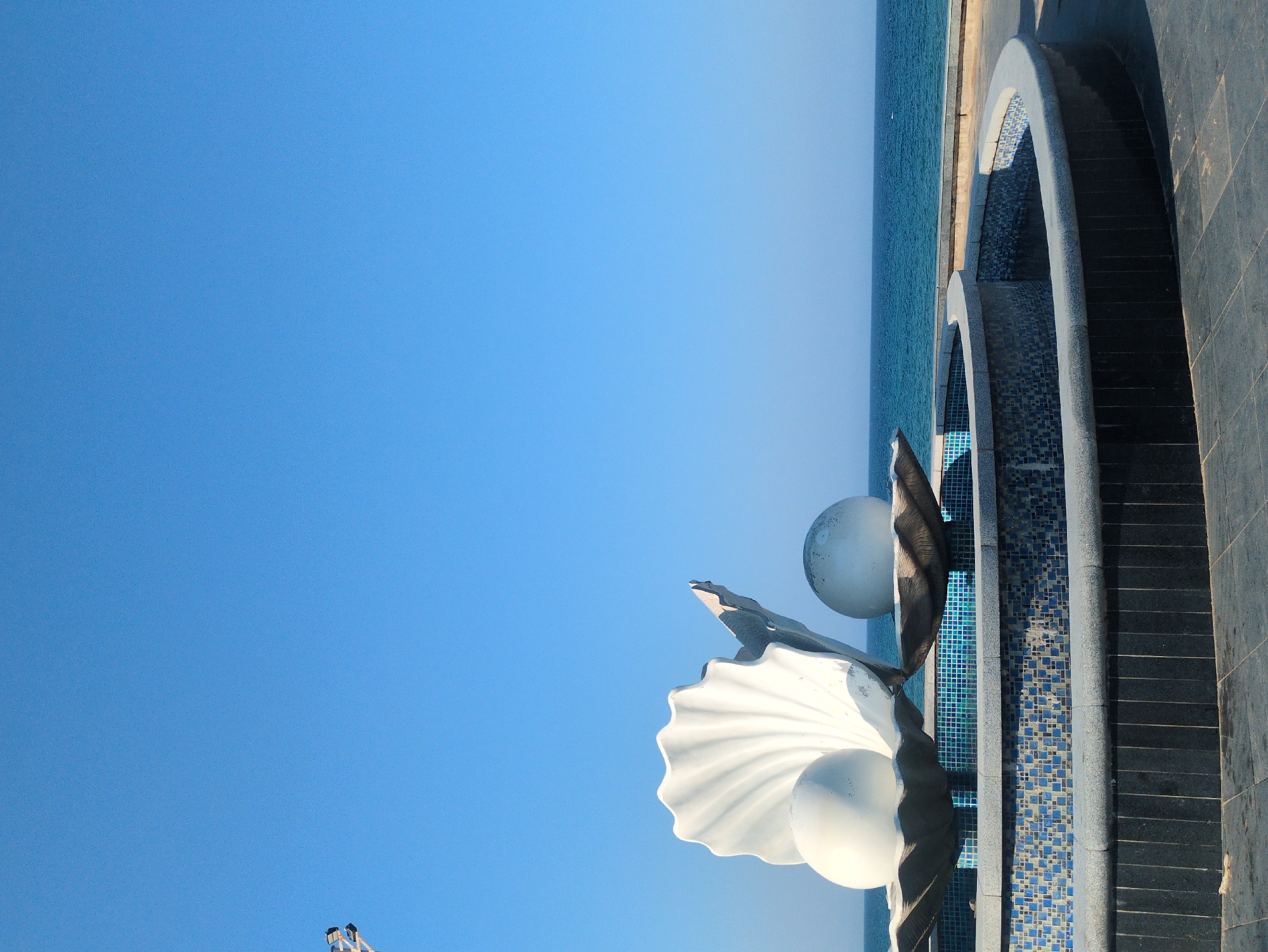The image shows a seaside structure in Tuy Hòa, Vietnam. The foreground features a large, decorative seashell sculpture with two opaque, white spheres embedded within. Adjacent to the sculpture is a fountain-like structure, lined with dark gray tiles and divided into sections with blue and white mosaic tile bottoms. Beyond the structure is a clear, light blue sky and a stretch of blue ocean.