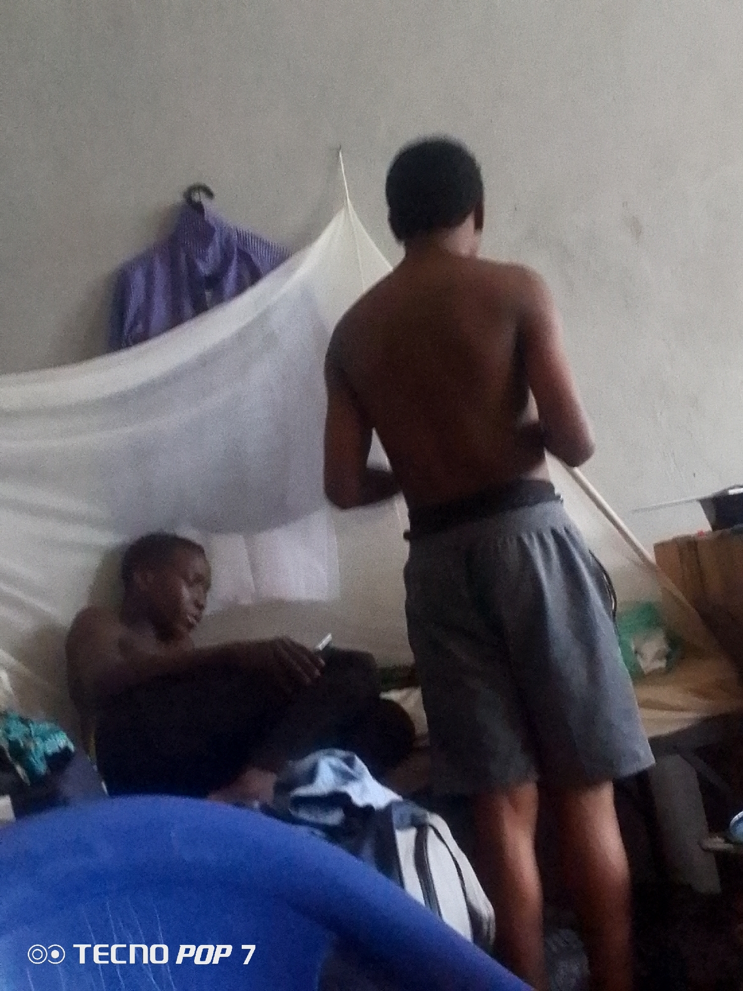 The image depicts an indoor scene with two young individuals. One person is seated, leaning back and holding a cigarette. This individual is wearing dark clothing. The other person is standing, facing away from the camera, and appears to be wearing grey shorts. Both individuals are shirtless.
The setting seems to be a room, possibly a bedroom, with a mosquito net draped above and behind the seated person. A purple and white striped shirt hangs on a hanger on the wall in the background. Various objects are scattered around the room, including what appear to be bags and bedding. The lighting suggests it is daytime, though the indoor setting makes it difficult to determine the exact time of day or weather.
The overall atmosphere is casual and relaxed. The image is watermarked with "TECNO POP 7" in the bottom left corner. The location context provided is Ipetumodu, Nigeria.