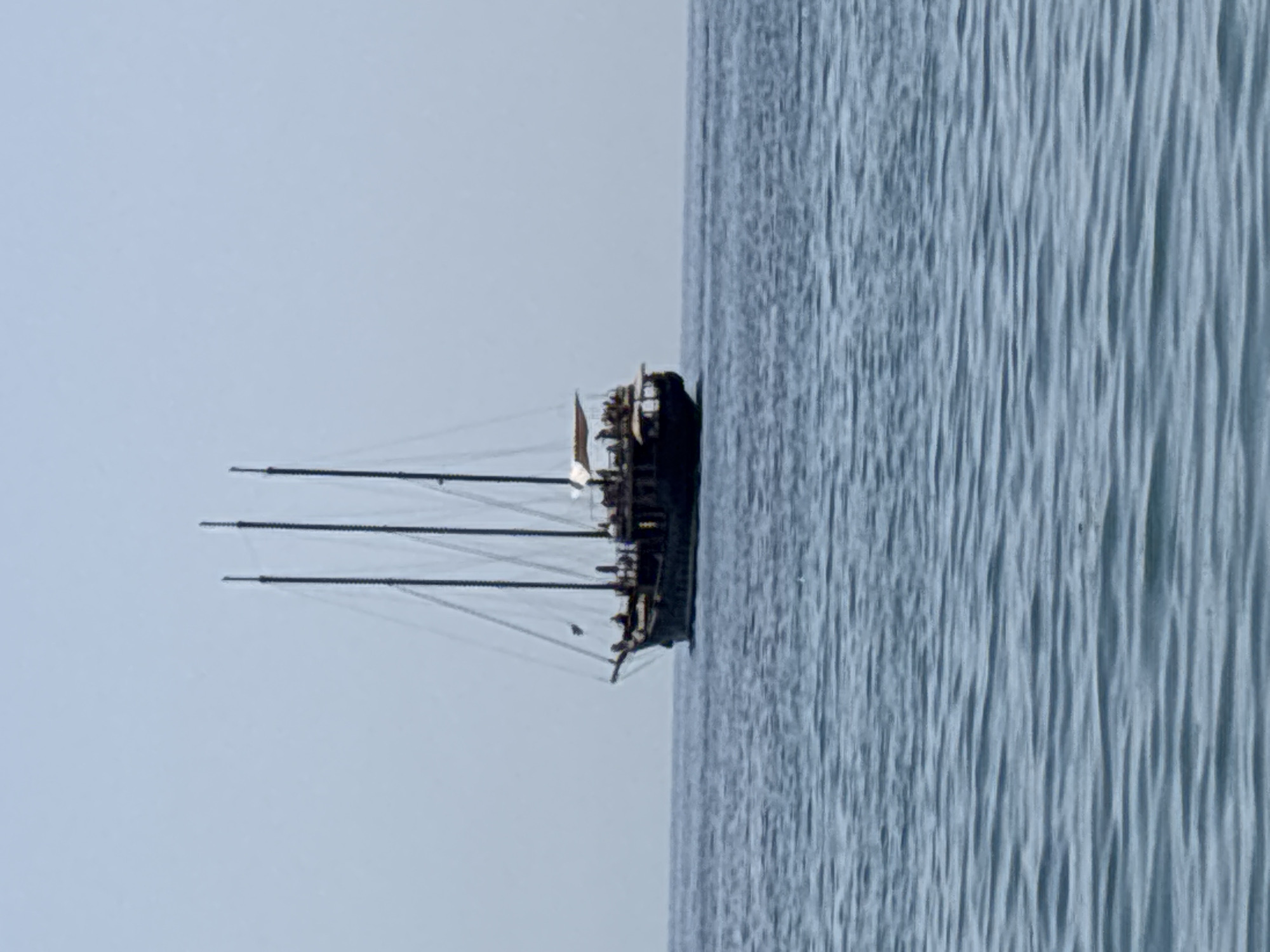 A dark-hulled sailboat with three masts is sailing on a calm, light blue sea. The sailboat is positioned in the center-right of the frame. It appears to be a tour boat, with a canopy covering the rear deck where several people are visible. The sky is a pale, hazy blue. The water is rippled, suggesting a gentle breeze. The image depicts a serene maritime scene likely near El Parador de las Hortichuelas, Spain, given the characteristic clear, calm waters of the Mediterranean coast.