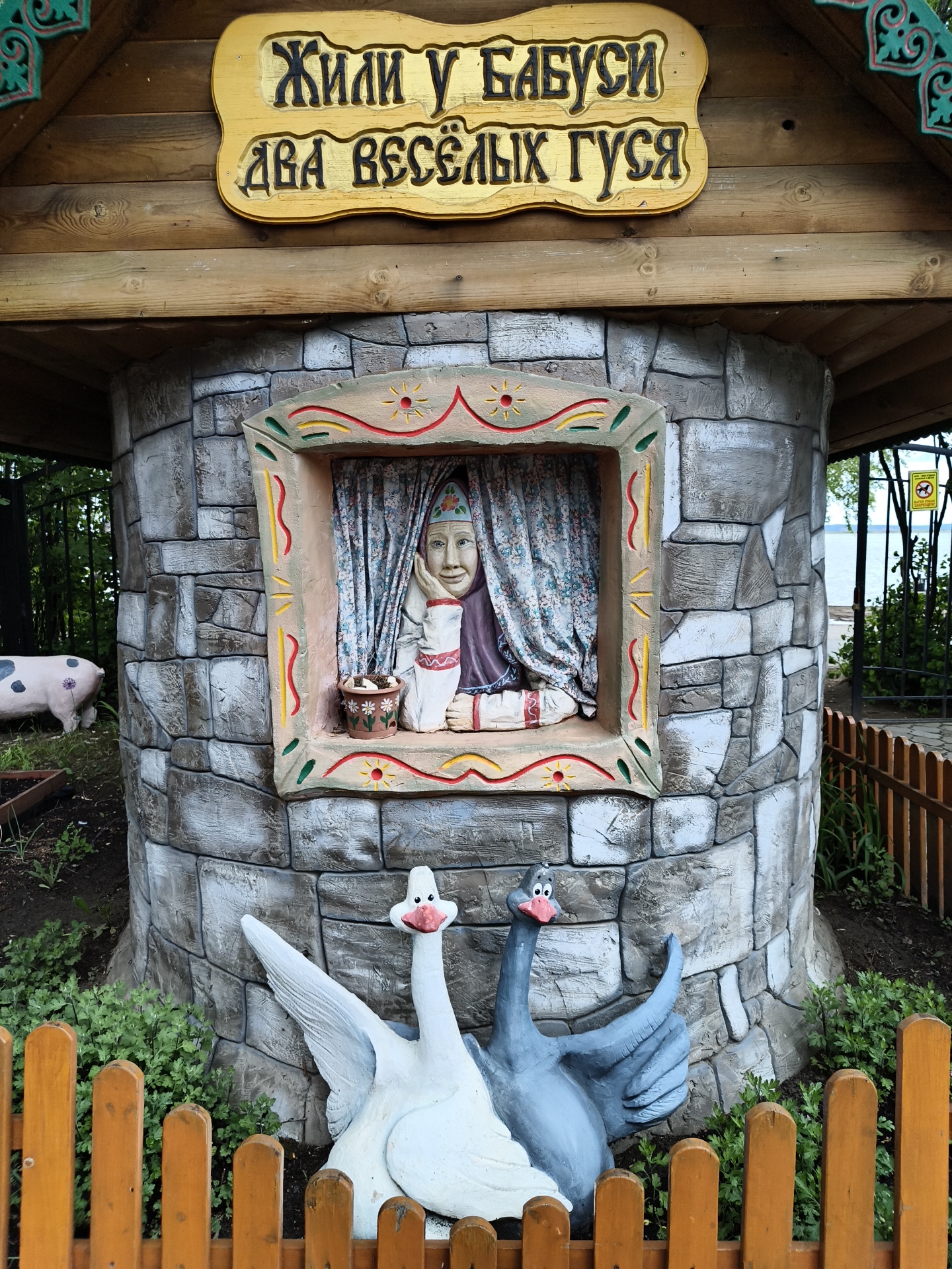 This image shows a sculpture of an old woman sitting in a window frame, with two geese in front of it. The sculpture is part of a public art installation located in the city of Chaikovsky, Russia. The sculpture sits under a wooden roof that has a sign, in Russian, that reads, "Жили у бабуси два весёлых гуся" (or "There lived a grandmother who had two merry geese"). The structure is built to look like stone with a rectangular window with a painted woman behind a curtain and a flower pot. The two geese are painted white and grey. A pig sculpture is nearby, in the foliage. A wooden fence surrounds the base of the sculpture.