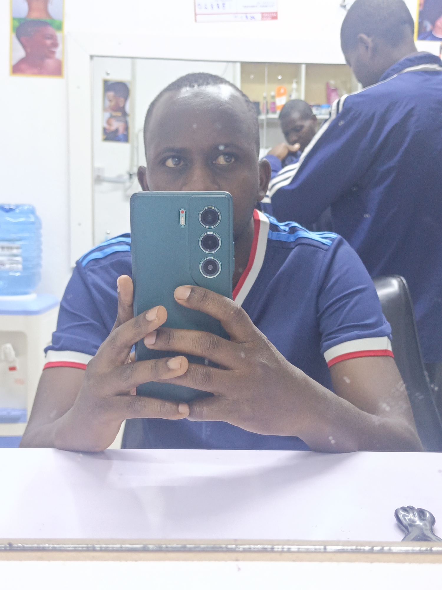 A man is taking a selfie in a mirror, likely in a barber shop or salon, as evidenced by other people in the background and posters on the wall. The man is wearing a blue t-shirt with white and red trim. He is holding a green smartphone up to his face. Behind him and to the right is another man in a blue jacket with white stripes. In the background on the left is a water dispenser. The man appears to be in a commercial establishment, perhaps in Nairobi, Kenya.