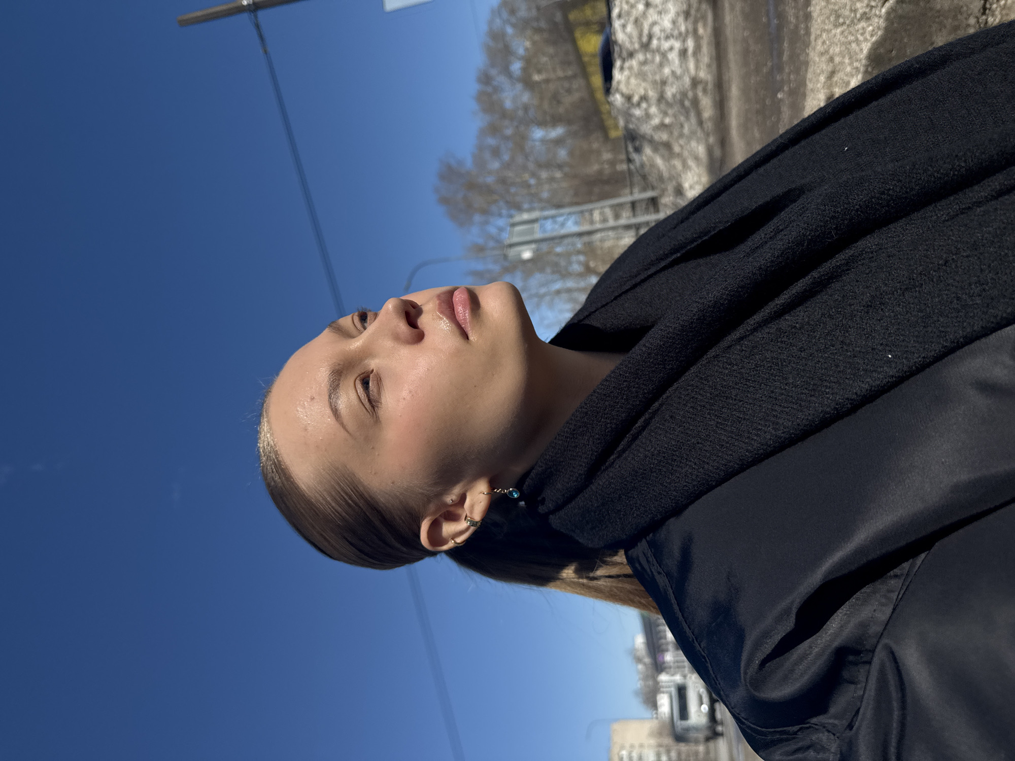 The image shows a woman from a low angle perspective, looking upwards. She is wearing a black jacket and a black scarf. Her hair is light brown and pulled back. She has multiple earrings in her left ear. The sky is a clear blue. In the background, out of focus, are trees, buildings, and street lights. Based on the available visual data, the location in Chaikovsky, Russia cannot be forensically confirmed.