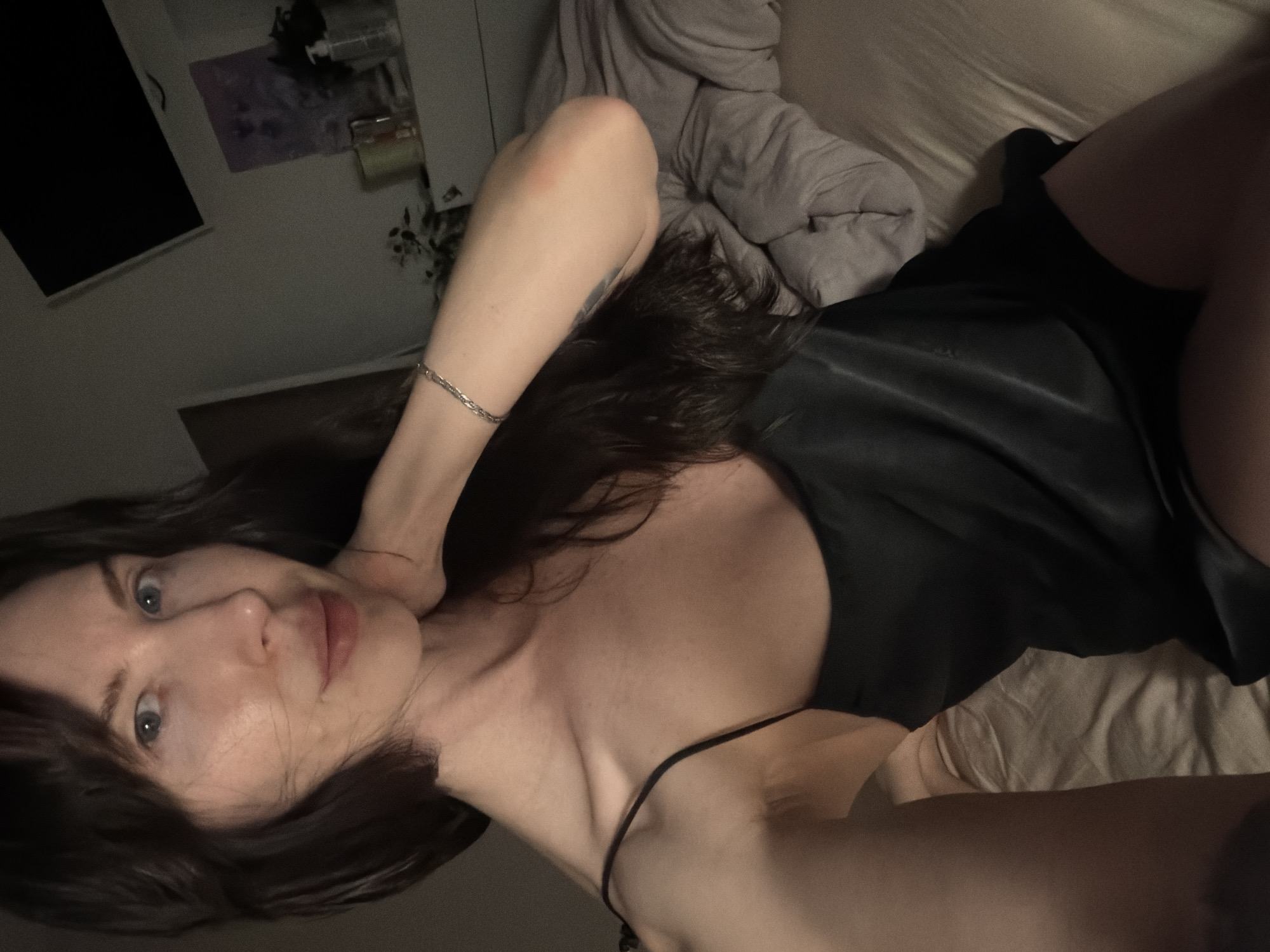 A woman lies on a bed, wearing a black satin-like dress with a thin strap visible on her shoulder. She has dark hair and light skin, with blue eyes visible in the close-up of her face. Her left arm is raised, and a silver bracelet is visible on her wrist. The background shows a dimly lit room, with a bed and a wall with a dark object, possibly a television, and other items on a shelf. The lighting suggests an indoor setting, possibly a bedroom. The location in the city of Perm, Russia, cannot be confirmed from the visual data.