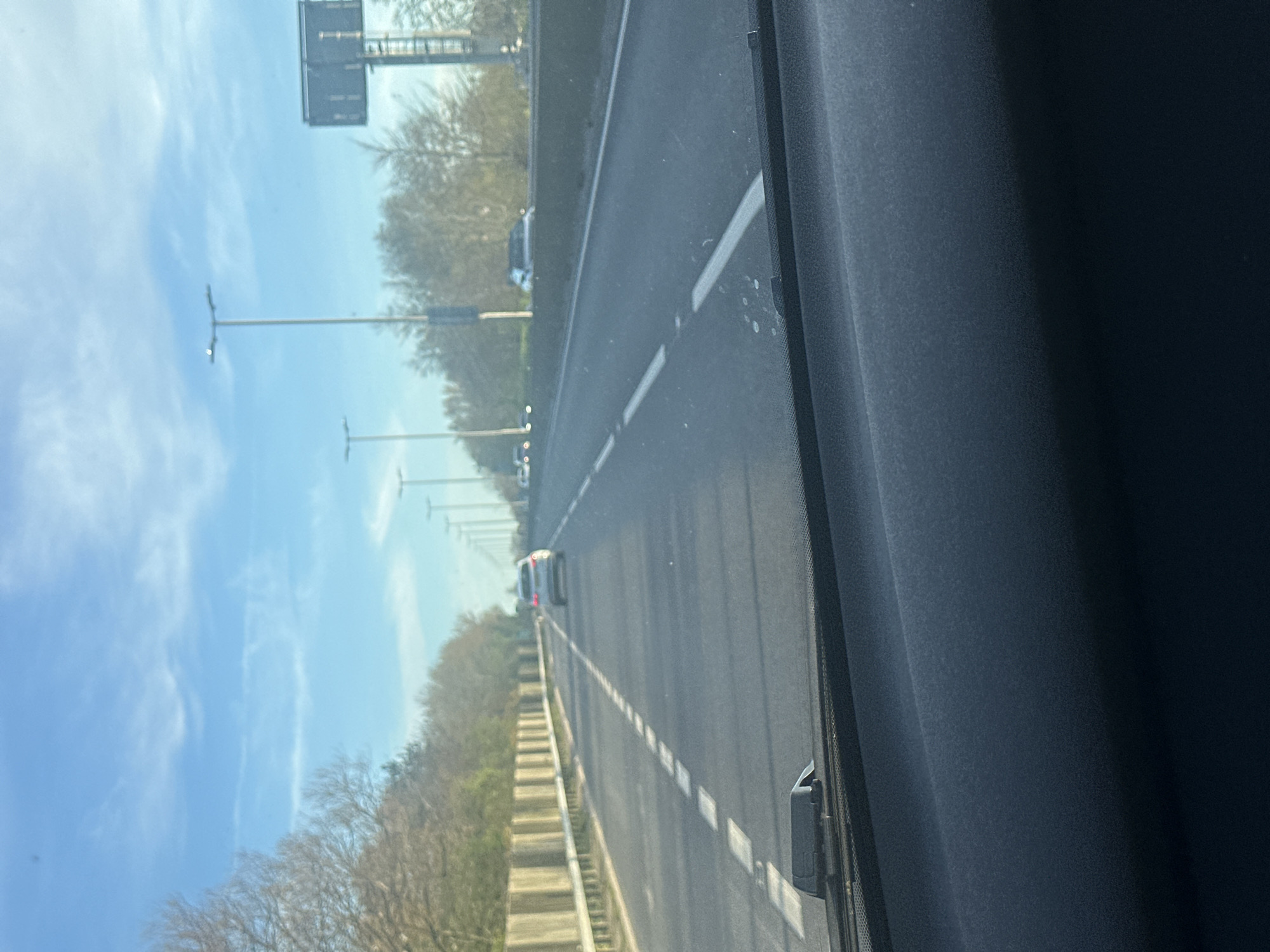The image depicts a highway scene from the interior of a vehicle, likely a car. The view is through the windshield, focusing on the road ahead. The road is multi-lane, with dashed white lines indicating lane divisions. Other vehicles are visible in the distance. To the left of the road is a shoulder area with a guardrail, beyond which are trees and a blue sky with some cloud cover. Streetlights are visible alongside the road. The interior of the vehicle is visible on the right side of the frame. Based on the road signs and overall environment, it is possible that the location is near Gorleston-on-Sea, United Kingdom.