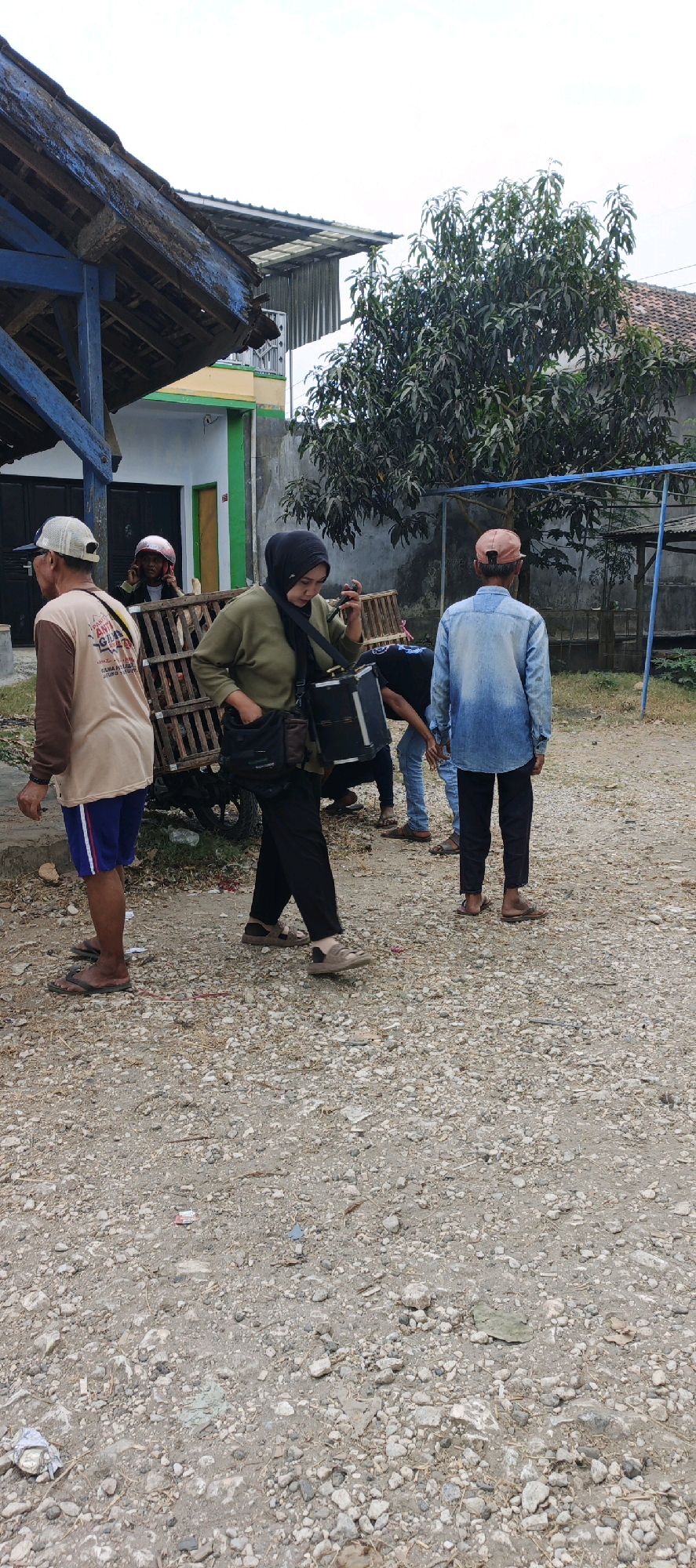 The image depicts an outdoor scene in Kasiyan, Indonesia, featuring several individuals interacting near a wooden structure and a cart. The environment is characterized by a gravelly ground with scattered debris.
In the foreground, a person wearing a tan long-sleeved shirt with brown sleeves, blue and white shorts, and sandals is facing away from the camera, looking towards the left. To their right, a woman wearing a hijab and olive-green long-sleeved shirt, black pants, and sandals is holding a black rectangular device with a strap over her shoulder. She appears to be looking at the device. Behind her, a wooden cart with a metal wheel is visible, loaded with what appears to be a cage.
Further back, another individual wearing a blue denim shirt and a light-colored baseball cap is facing away from the camera, to the right of the woman with the hijab. A person in a light blue denim shirt and dark pants is partially visible to the right of the woman with the hijab. In the background, a person in a red helmet is seated, looking to the left, possibly on or near the cart. A building with a blue-painted wooden support structure and a weathered roof is on the left. A tree with dark green foliage stands in the middle-right of the image, with a fence visible behind it. The sky is overcast and light in color.
