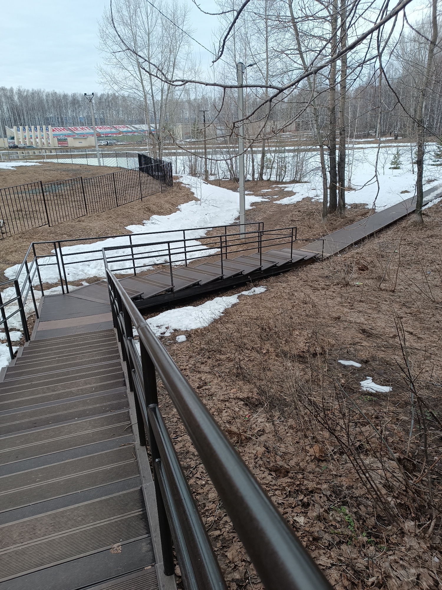 The image depicts an outdoor scene in what appears to be a park or recreational area. There are metal stairs with wooden treads and a dark metal railing in the foreground, leading down a slope covered with fallen leaves and patches of melting snow. To the right of the stairs is a wooden boardwalk or path winding through the trees. The ground is covered with brown leaves, and there are bare trees and bushes scattered throughout the scene. In the background, there is a fenced area and a large building with stadium seating, possibly a sports complex. Based on the context provided, the location is likely within the city of Chaikovsky, Russia. The overcast sky suggests it is a cloudy day.