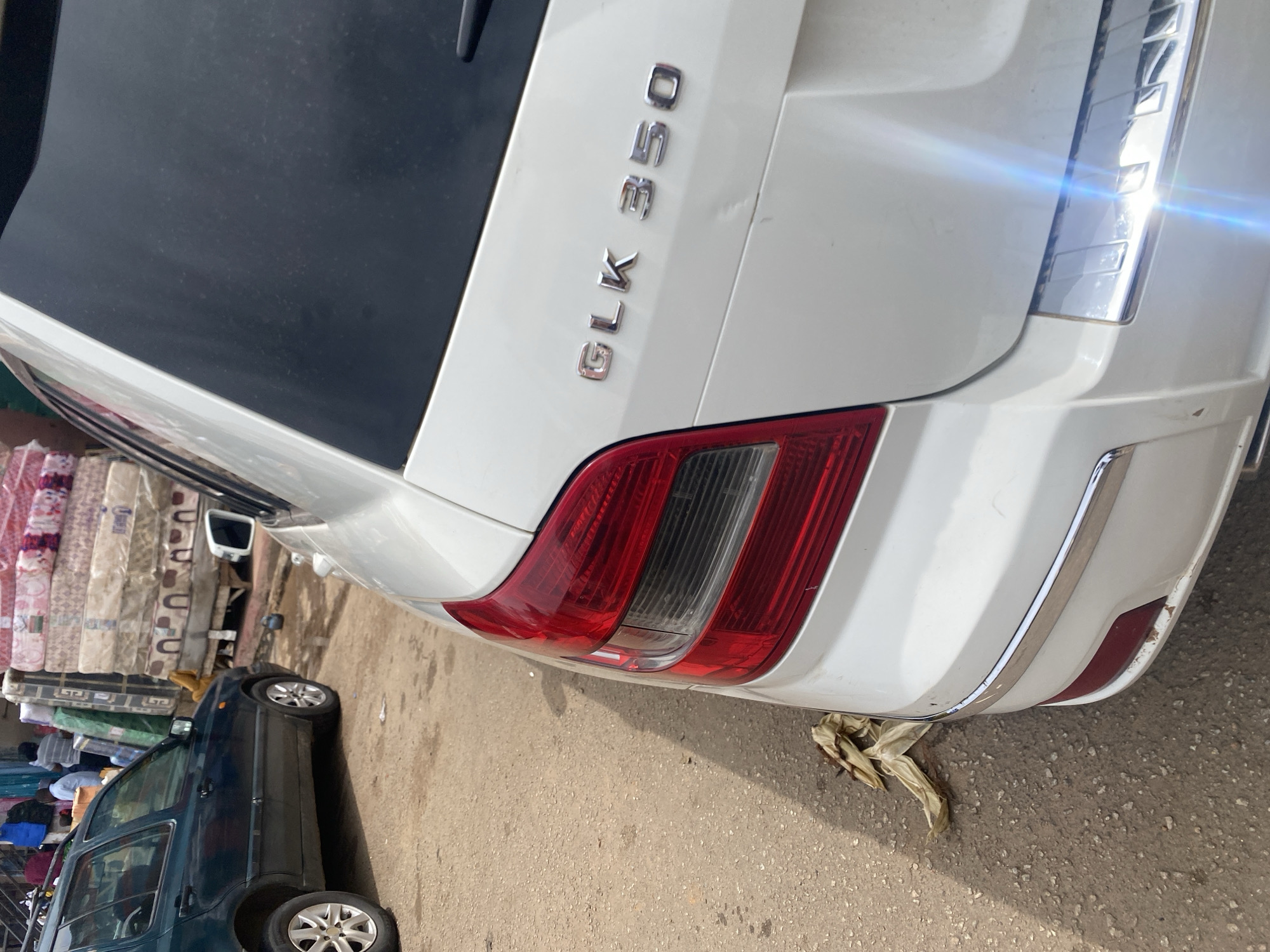 The image captures a close-up view of the rear of a white Mercedes-Benz GLK 350. The car is parked on a dusty, paved street in Jos, Nigeria, evident from the context. To the left of the car, there is a display of mattresses, suggesting a commercial area or market. The lighting indicates it is daytime, with the sun casting reflections on the car's metallic trim and taillights. The overall atmosphere appears to be an outdoor street scene, likely in a bustling commercial district. The visible text on the car reads "GLK 350", identifying the vehicle's model.