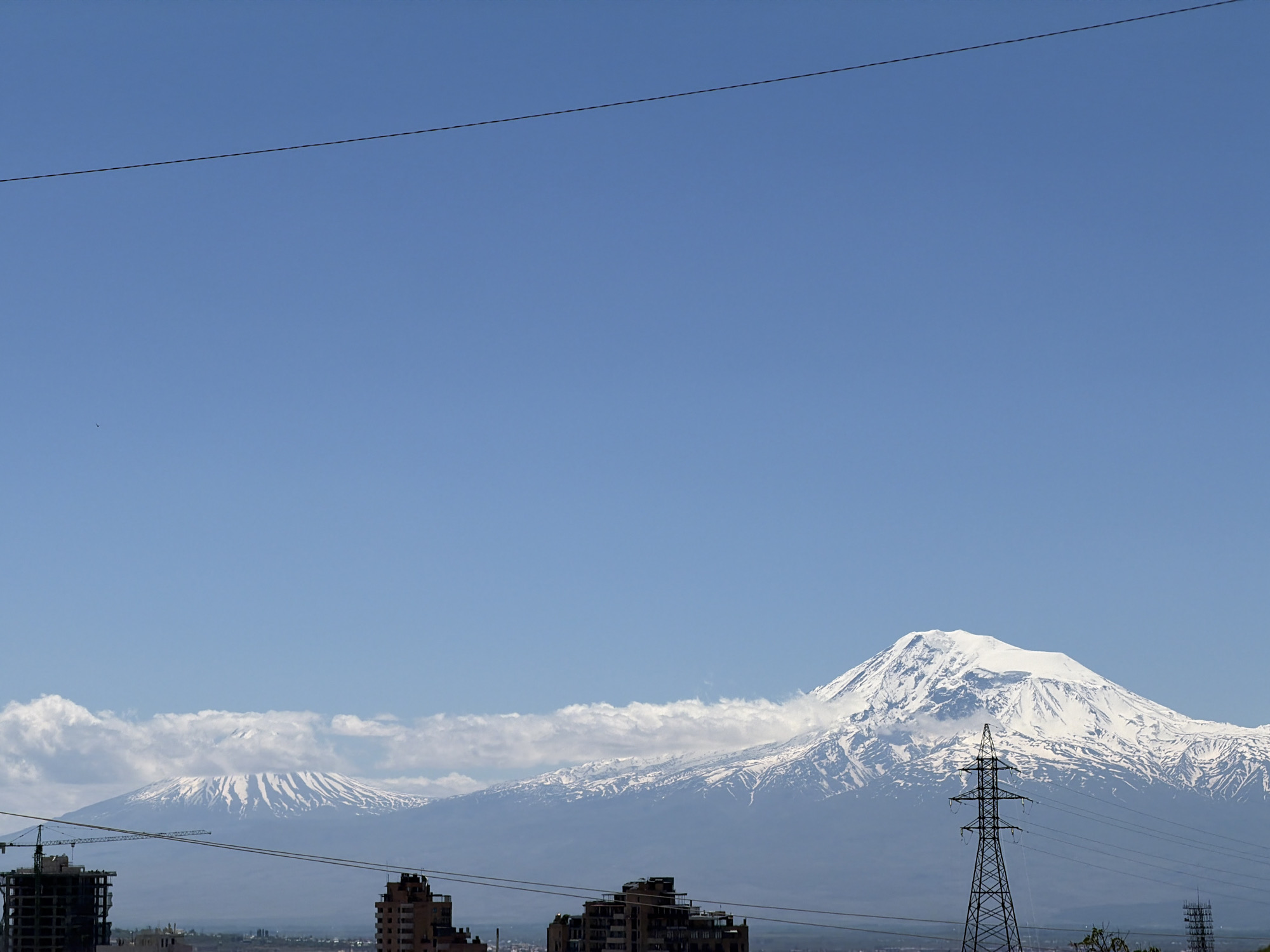 The image shows a clear, blue sky above a cityscape with mountains in the background. In the foreground, there are buildings under construction and completed apartment blocks, along with a power transmission tower. Two large, snow-covered mountains dominate the horizon. The scene is likely taken in Yerevan, Armenia, given the distinctive appearance of Mount Ararat in the background. There are no people visible in the image.