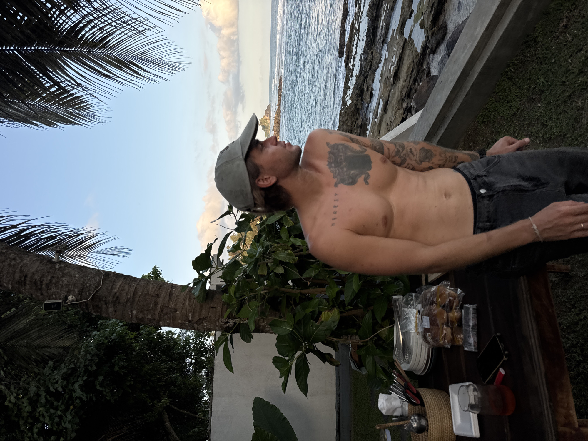 A shirtless man wearing a grey baseball cap and dark grey jeans stands on a grassy area next to a wooden table. The man has tattoos on his chest and arms. On the table are plates, cutlery, a glass of red liquid, a smartphone, and a package of buns. The table is situated on a patio overlooking a rocky coastline and the ocean. Palm trees are visible in the background, with the sky showing a few clouds. The scene is located in Goviyapana, Sri Lanka.