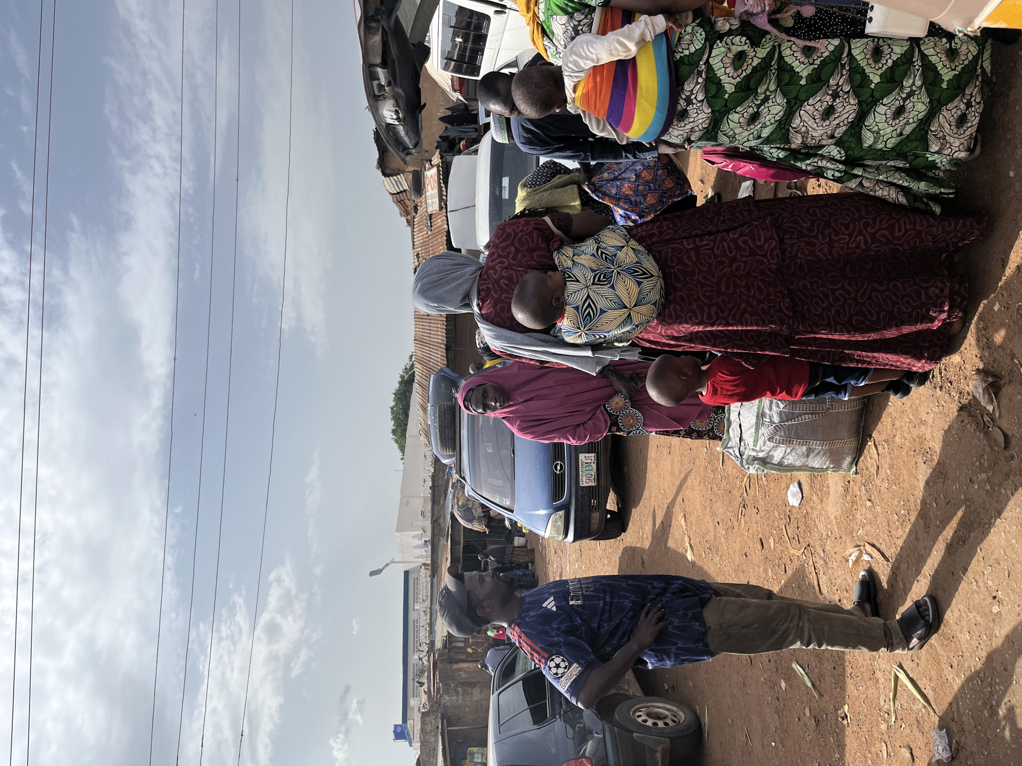 In Rafin Par, Nigeria, an outdoor scene unfolds with several people and vehicles under a partly cloudy sky. A man wearing a dark blue soccer jersey with white accents and khaki pants stands to the left, facing right and looking away from the camera. Next to him, to the right, a woman in a vibrant pink headscarf and dress stands with her hands clasped. Behind them, a blue minivan is parked, its license plate clearly visible. Further back, more people and vehicles are present, including what appears to be a white van and other structures that suggest a market or busy thoroughfare. Two women are carrying babies on their backs using colorful wraps. One baby is wrapped in a pattern of blue, yellow, and white, while the other is in a rainbow-striped wrap. A young child in a red shirt and blue shorts stands near the women, looking towards the right. A large, grey woven bag sits on the ground between the child and the women. The ground is covered in reddish-brown dirt, with scattered leaves and debris. Buildings with corrugated metal roofs are visible in the background, along with a white building and a tower with lights. Power lines are strung across the top of the frame. The overall impression is one of a bustling, everyday scene in a Nigerian town.