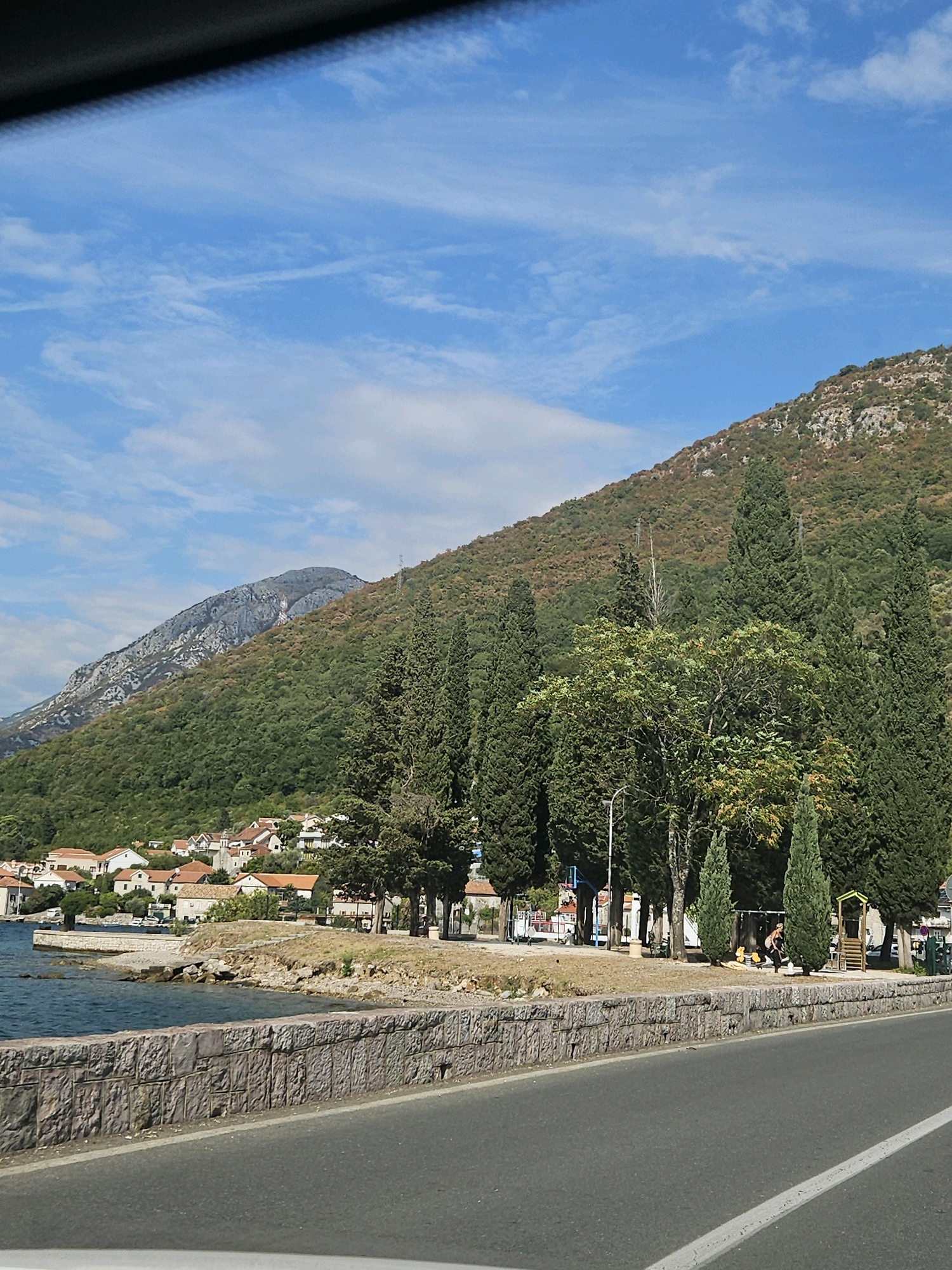 The image captures a coastal road scene with a backdrop of lush green mountains and a clear blue sky dotted with white clouds. In the foreground, an asphalt road curves to the right, bordered by a textured stone wall. Beyond the wall, a rocky shoreline meets the calm, blue water of the bay. A village with terracotta-tiled roofs is nestled along the coast, appearing as a collection of white buildings. Tall, slender cypress trees and broader leafy trees line the waterfront, creating a natural screen between the village and the road. A small playground area is visible with a swing set and a yellow play structure. A person is seen interacting with the playground equipment. The mountains in the background are densely forested with patches of exposed rock.