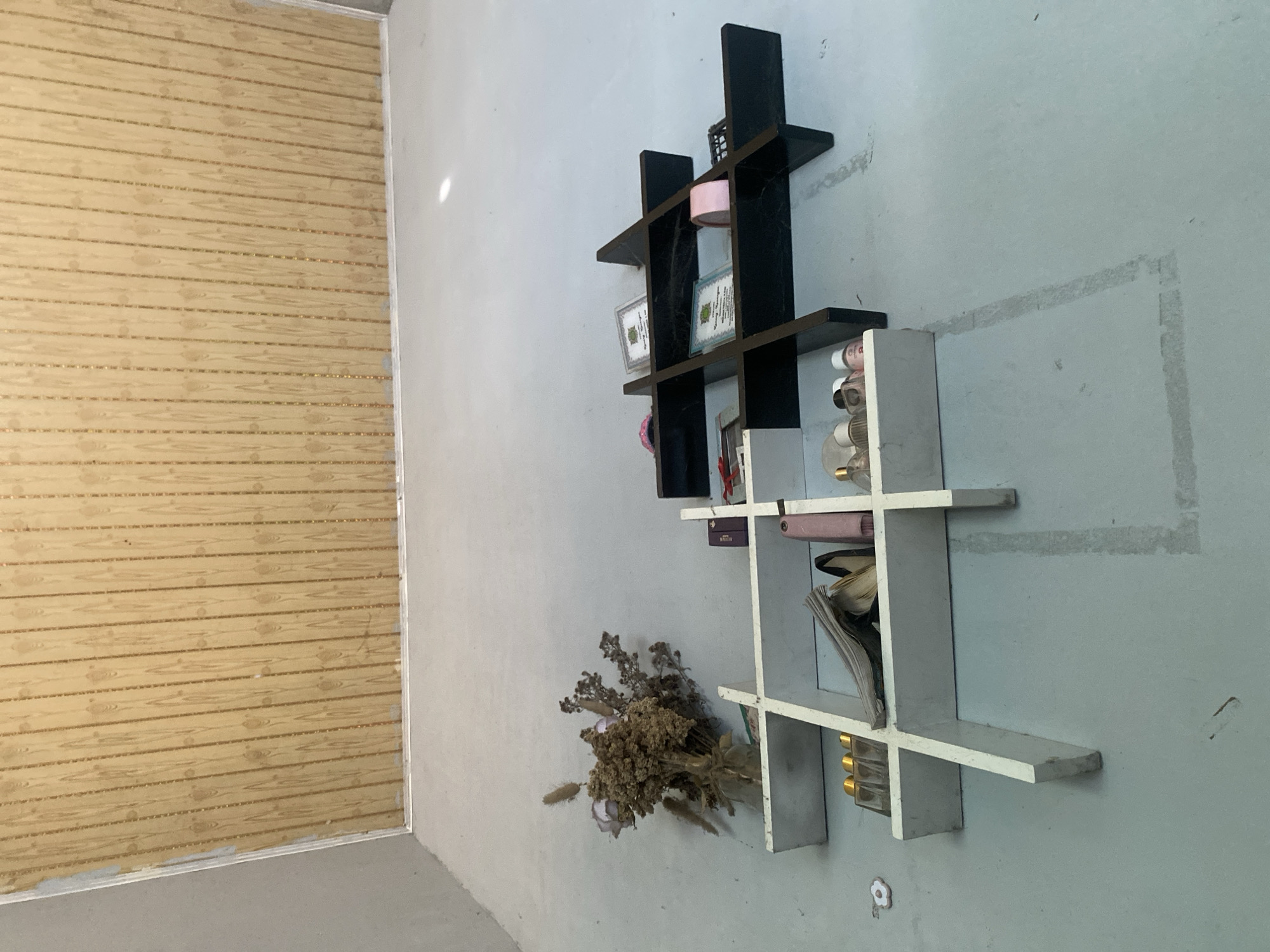 The image depicts an interior scene with a light blue, concrete floor and a light-colored wall in the background that transitions into a wood-paneled wall at the top. In the foreground, there are two white shelves and one black shelf unit.
The white shelf unit on the left is L-shaped and has several compartments. It holds a bouquet of dried flowers in a glass container, various small bottles with golden caps, a stack of papers, and a pink cylindrical object.
The black shelf unit is a tic-tac-toe design and is placed to the right of the white shelf. It contains two framed certificates or documents with green emblems, a pink roll of tape, and a black object that appears to be an electronic device.
The lighting in the scene is diffuse, with some reflections visible on the floor. The overall impression is of an organized display of items on shelving units in what appears to be a room under renovation or in a state of disuse. There are no people visible in the image. The location is identified as Meunasahlhok, Indonesia.