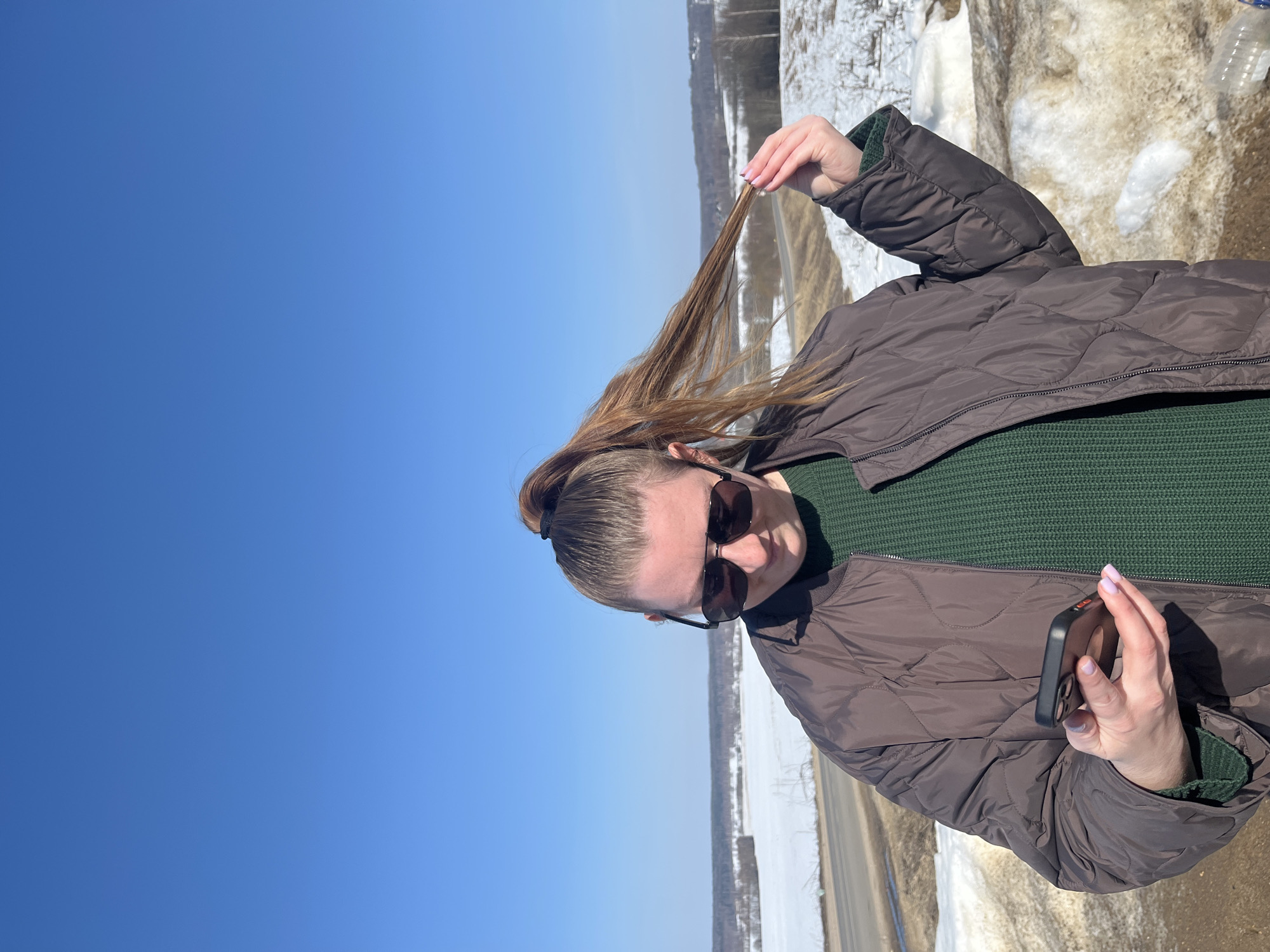 A female individual is depicted outdoors, in the city of Zubovo, Russia, under a clear blue sky. She wears dark sunglasses, a dark brown quilted jacket, and a dark green knit sweater. Her light brown hair is styled in a high ponytail, with her right hand grasping a section of it. She holds a dark-colored smartphone in her left hand, her gaze directed downwards towards the device. The background features a wide, open landscape partially covered in snow, with a two-lane asphalt road curving through the middle ground. Distant trees and a horizon line are visible. Patches of melting snow and dirt are present in the immediate foreground.