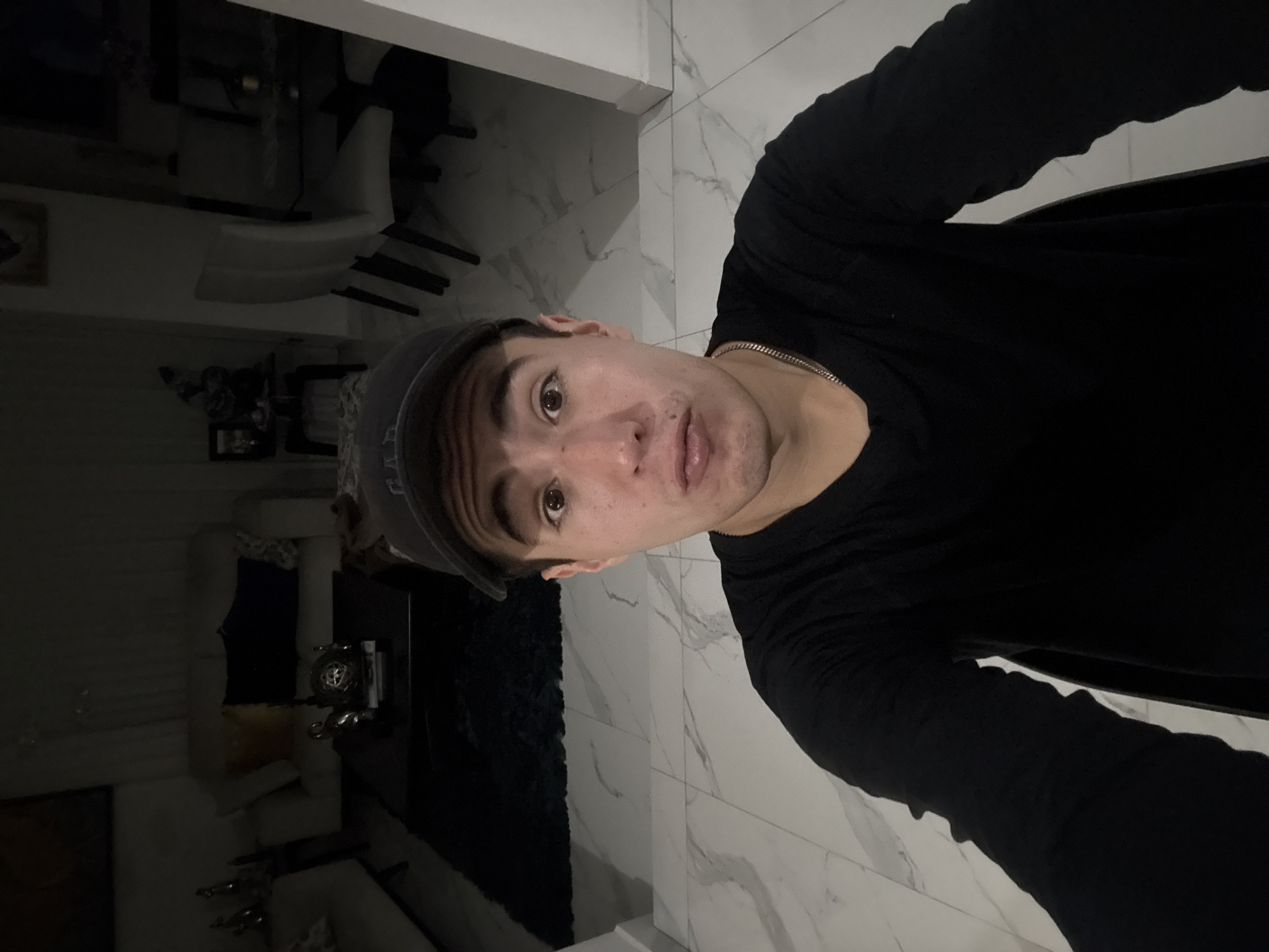 A man is standing indoors, wearing a dark long-sleeved shirt and a baseball cap. The background includes a white tiled floor and furniture that is partly obscured by shadow. The location cannot be definitively identified as San Bernardino Tlaxcalancingo, Mexico from the visual information provided.