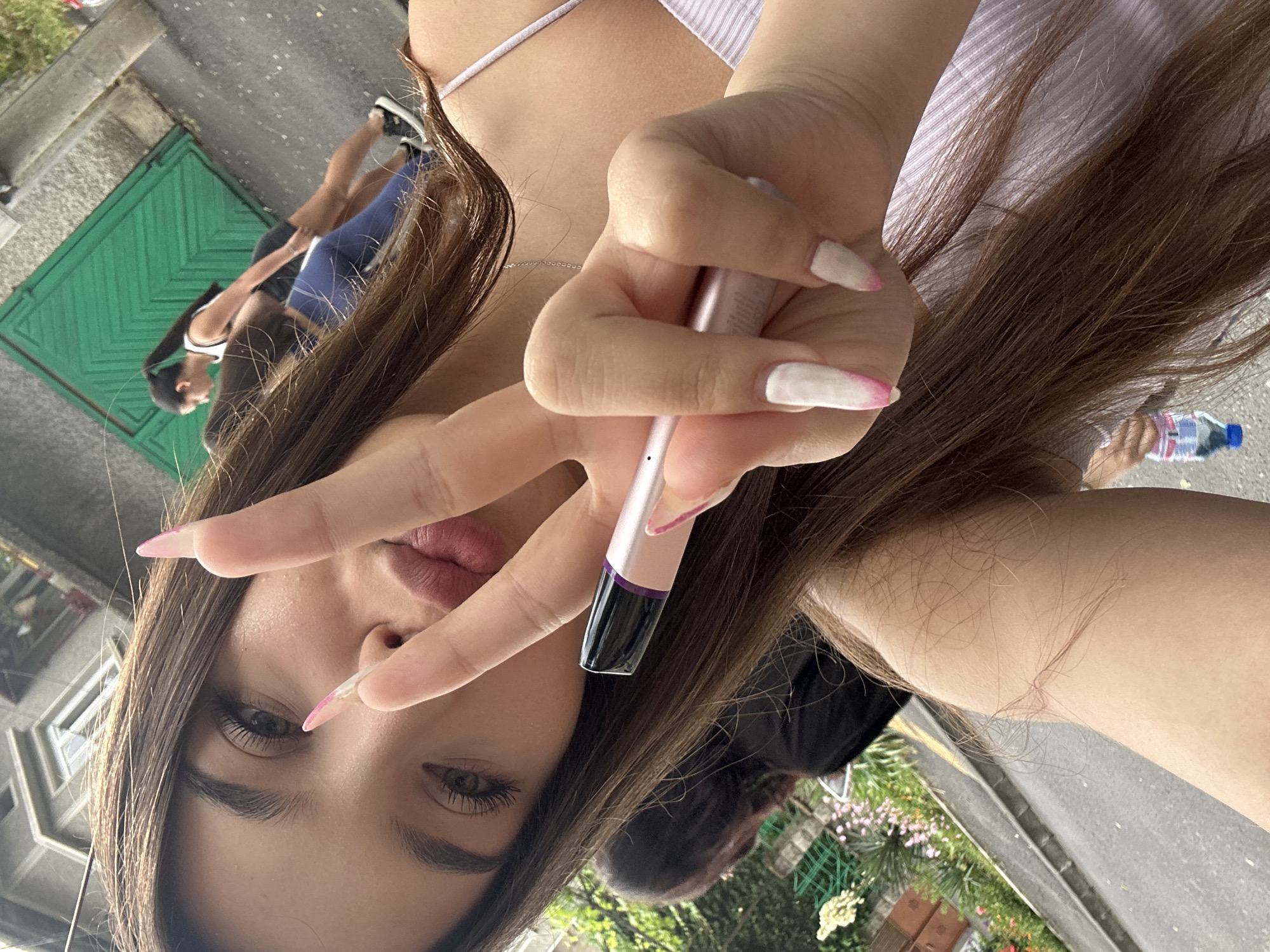 A young woman with long brown hair and long, manicured nails, painted white with pink tips, is holding a small, silver and purple electronic device, possibly a vape pen, in her right hand. She is making a peace sign with her index and middle fingers. Her lips are painted with a pink lipstick, and her eyes are green with long eyelashes. She is wearing a light purple top with thin straps. In the background, two other women are visible. One, wearing a white tank top and dark shorts, is walking away from the camera, her dark hair pulled into a ponytail. The other, wearing a black top, is seen from behind. The scene appears to be outdoors, with a paved walkway or road and some greenery, including flowering plants, on the left. A building with windows is visible in the upper left corner, and a green wooden door or gate is in the upper right. The overall lighting suggests it is daytime. The location is identified as Shumen, Bulgaria.