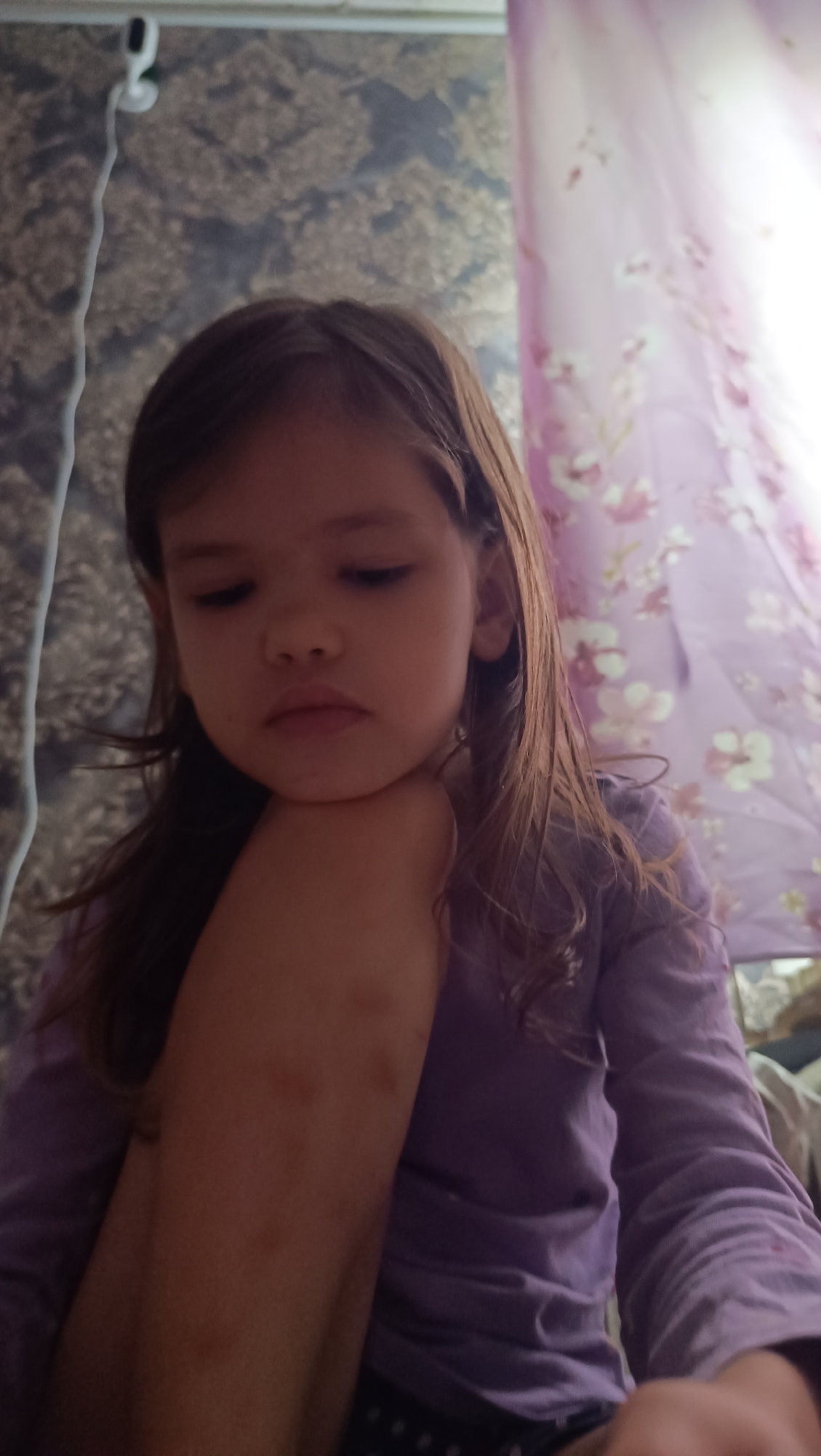A young girl with brown hair is captured in a close-up shot, her head resting on her arm. She is wearing a purple long-sleeved shirt. The background features a patterned wallpaper and a draped fabric with a floral design. The lighting suggests it is daytime. The girl's expression appears thoughtful or perhaps a little sad, with her eyes looking downwards. The scene seems to be indoors, possibly in a bedroom or living area. There are no visible signs of text in the image.