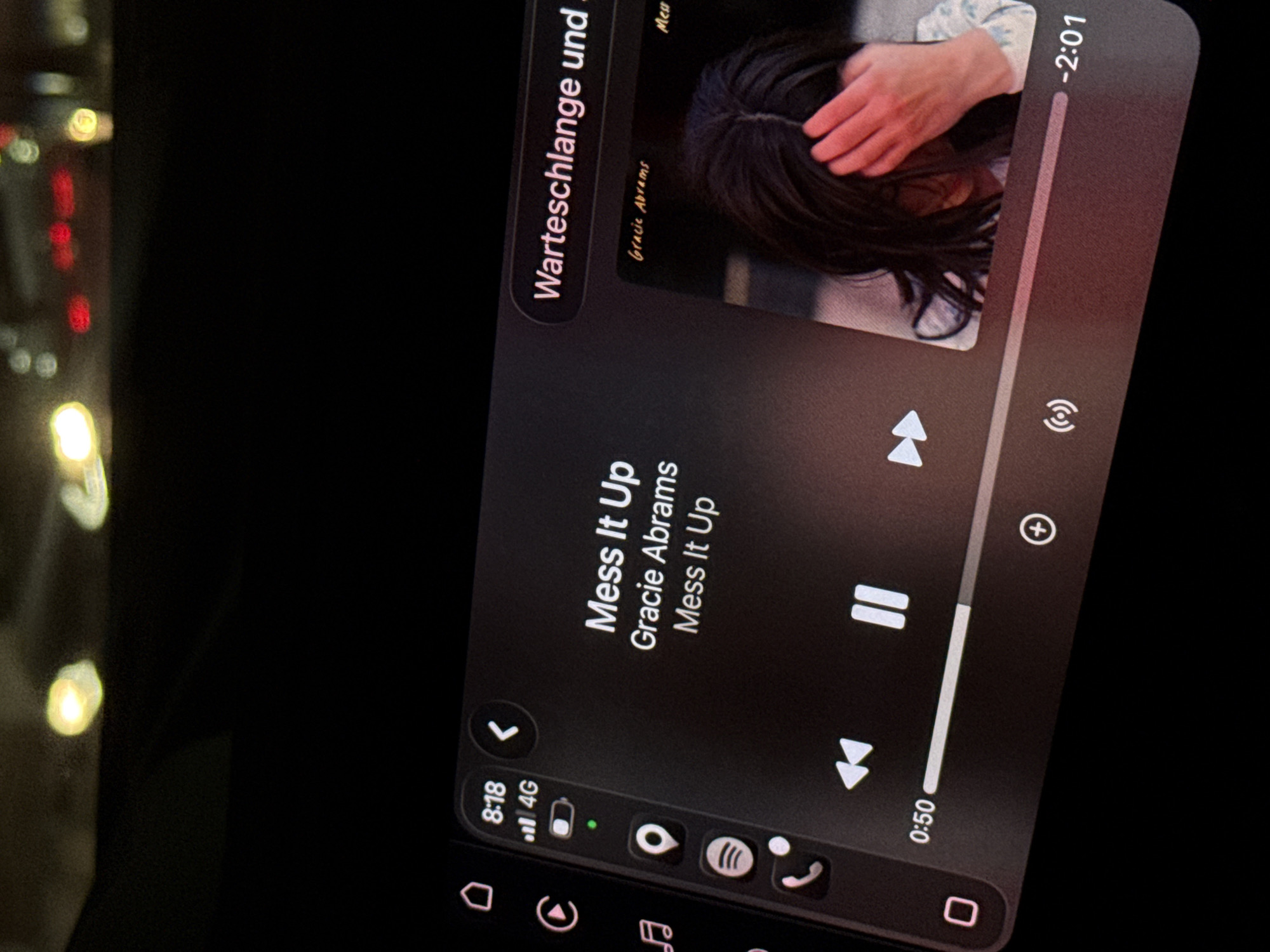 The image shows a digital display of a car's infotainment system. The screen displays the playback interface for a song titled "Mess It Up" by Gracie Abrams. The song is 50 seconds into its runtime, with 2 minutes and 1 second remaining. The user interface also shows standard icons, including mobile network signal strength (4G), battery level, location services, Spotify, and a phone call icon. The infotainment system appears to be a dark mode. The background is black and blurry.