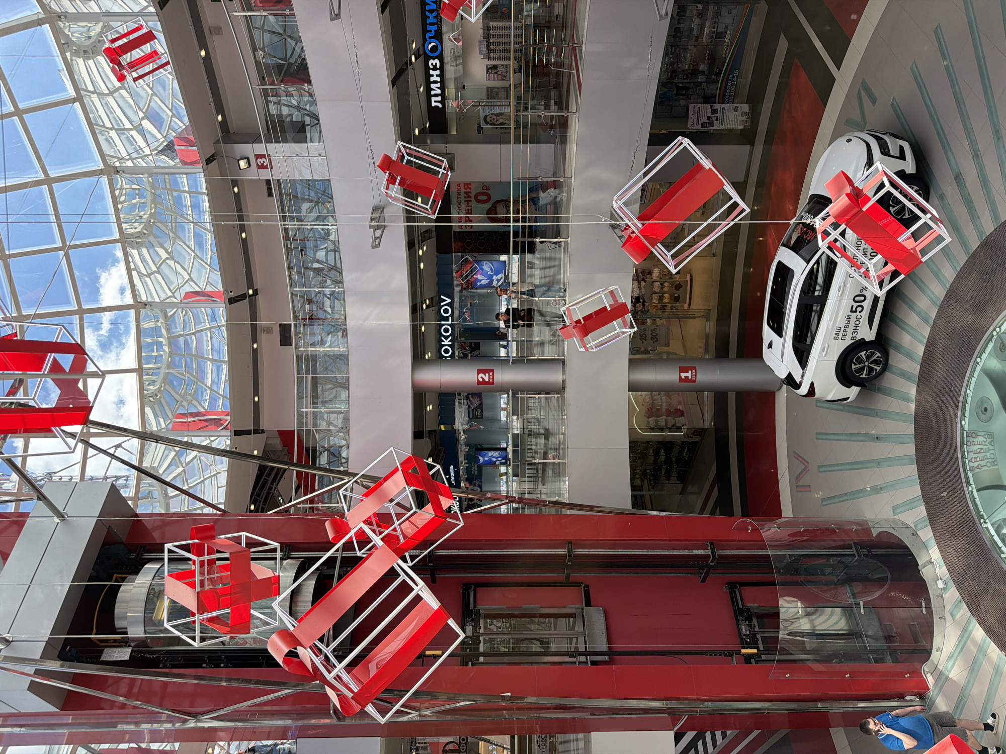 The image depicts the interior of a multi-level shopping mall in Izhevsk, Russia, viewed from an elevated position looking down. The architecture features a glass ceiling with a geometric pattern allowing natural light to illuminate the space. Red and white decorative structures resembling wrapped gifts are suspended throughout the atrium.
Several floors of retail space are visible, with signs for businesses such as "Linzochki" (optics store), "Sokolov" (jewelry), and offers for eye exams. A white car is positioned on the ground floor near a circular architectural feature, displaying a promotion of "50%" off the first installment. A man in a blue shirt and shorts is visible near the circular feature.