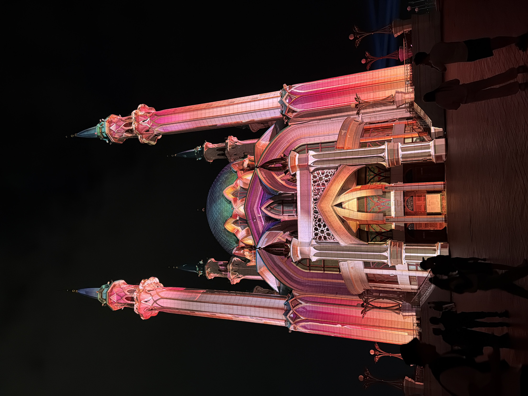 The image shows the Qolşärif Mosque in Kazan, Russia, illuminated at night. The mosque features tall minarets and a central dome, all lit with pink and white lights. Shadows of people are visible in the foreground, suggesting they are observing the mosque. The background is black.