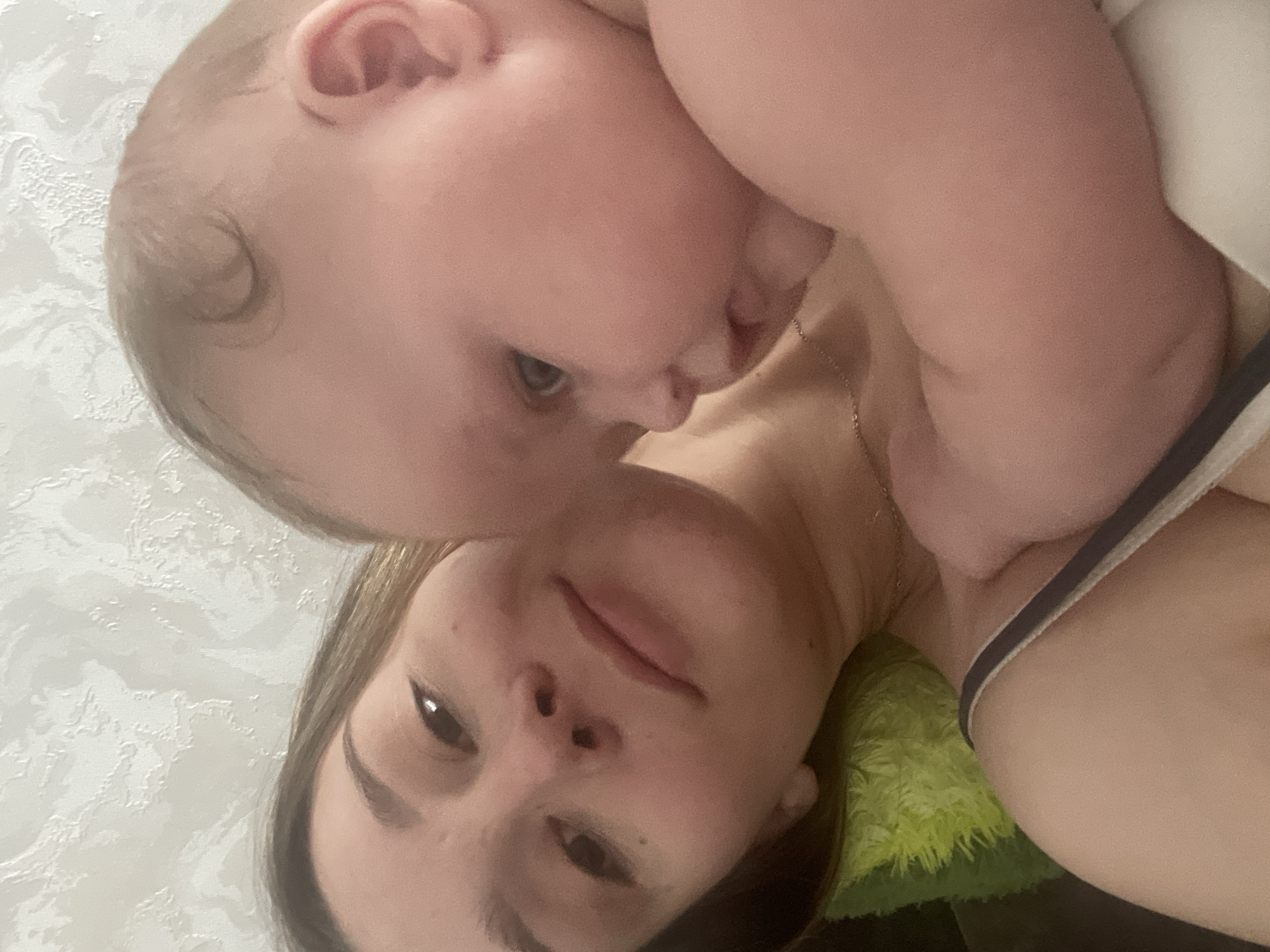 In the close-up selfie, a woman with light brown hair and brown eyes is holding a baby. The woman's head is tilted to the side, and she is smiling. The baby is facing away from the camera, with its head resting on the woman's shoulder. The background is a textured white wall. The image appears to be taken indoors.
