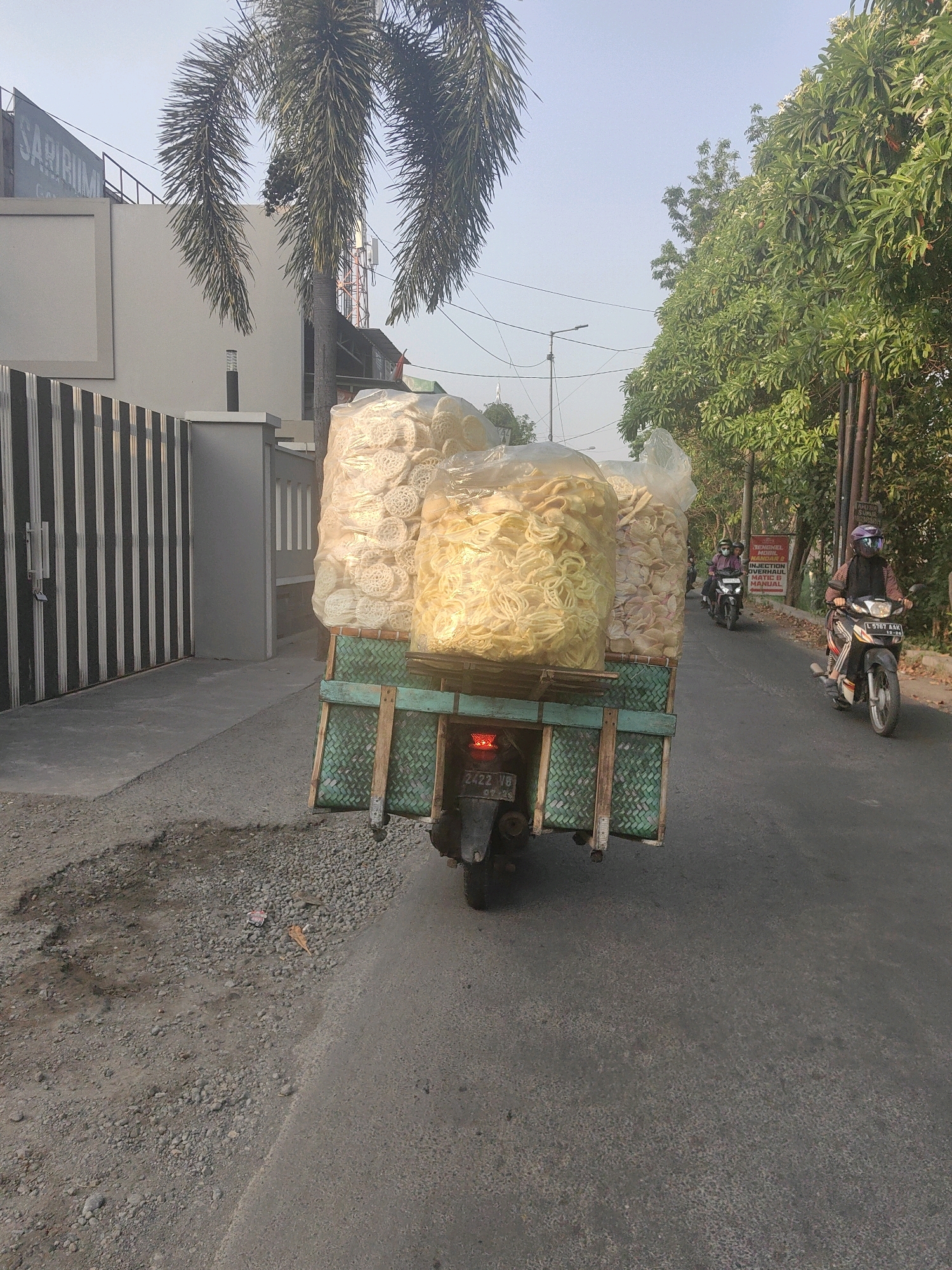 In Masangan Wetan, Indonesia, a scooter is transporting a large quantity of bagged snacks. The scooter, with the license plate "2422 VB", is overloaded with at least three large transparent plastic bags filled with light-colored, circular snacks, possibly crackers or chips. These bags are stacked high on a flatbed attached to the back of the scooter, which is secured by a woven mat material on its sides. The scooter is on an asphalt road, which appears to be in moderate condition with some gravel and debris on the shoulder. In the background, there are buildings, palm trees, and lush green foliage. Two other scooters are visible further down the road, also carrying passengers. The sky is a clear, pale blue.