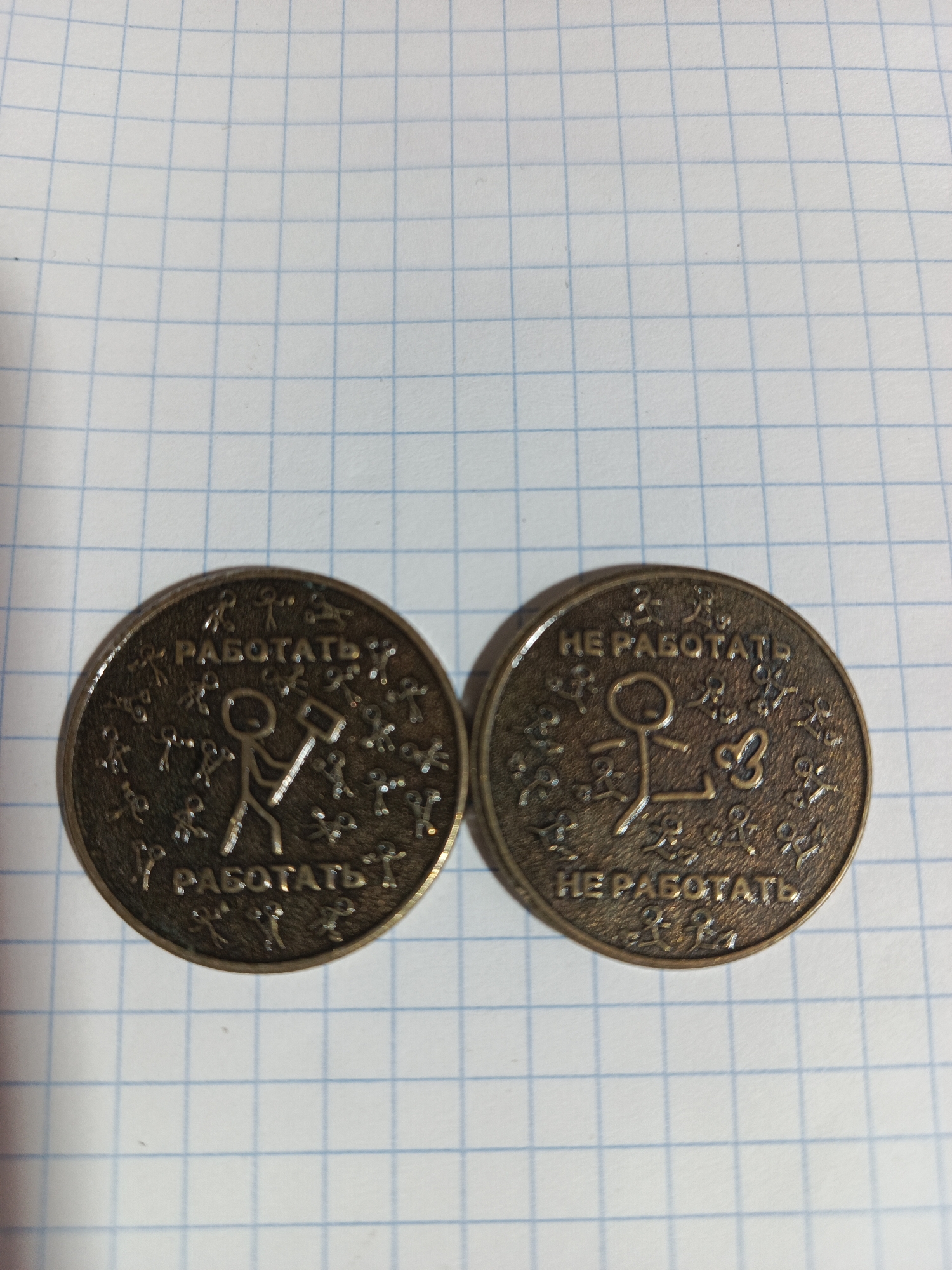 Two dark, circular metallic tokens rest side-by-side on a white sheet of graph paper. The paper features a grid of light blue lines.
The token on the left depicts a central stick figure holding a hammer, striking downwards. Surrounding this figure are numerous smaller, stylized stick figures or symbols. Along the top edge, the text "РАБОТАТЬ" (RABOTAT', meaning "TO WORK" in Russian) is inscribed, with the same text repeated along the bottom edge.
The token on the right features a central stick figure in a relaxed, possibly reclining, pose. A small butterfly symbol is positioned near its upper body. Similar to the other token, various smaller, stylized stick figures or symbols encircle the central design. The inscription along the top edge reads "НЕ РАБОТАТЬ" (NE RABOTAT', meaning "NOT TO WORK" in Russian), which is also repeated along the bottom edge.
Both tokens appear to have a slightly raised rim and a somewhat aged or antiqued patina, suggesting a brass or bronze material. The surface of both tokens is textured with small, irregular indentations, giving them a rustic appearance. The tokens are horizontally aligned, occupying the lower central portion of the image. The graph paper background provides no contextual information regarding any specific urban or outdoor location.