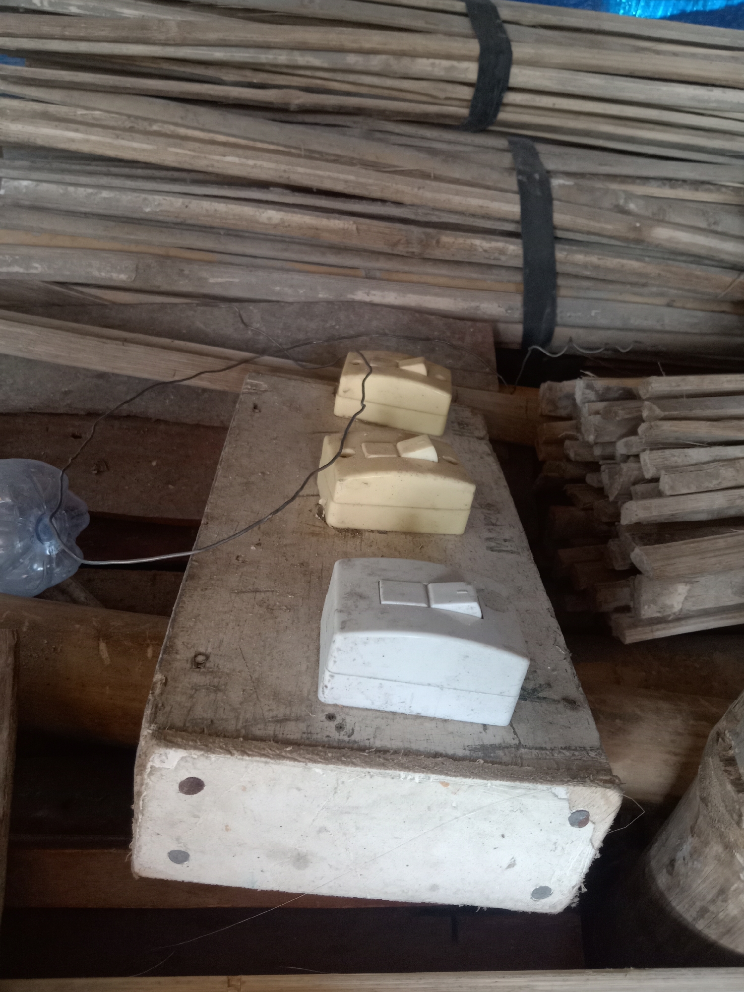 The image depicts an indoor scene, likely a storage area, characterized by a clutter of wooden materials and electrical components. In the foreground, a white electrical switch plate with two toggle switches is positioned on a rough wooden plank. Two smaller, beige electrical outlets are placed behind it on the same plank. A clear plastic bottle with some liquid is visible to the left, with a thin wire extending from it and connecting to the electrical components. The plank itself rests on other wooden structures. In the background, stacks of long, thin wooden strips are bundled together with black straps, and further to the right, more bundles of wood are visible. The overall environment appears somewhat unorganized and utilitarian, suggesting a workshop or storage space. The specific location in Stiaji, Indonesia, is not visually evident from the image itself, but the presence of common building and electrical materials is apparent.