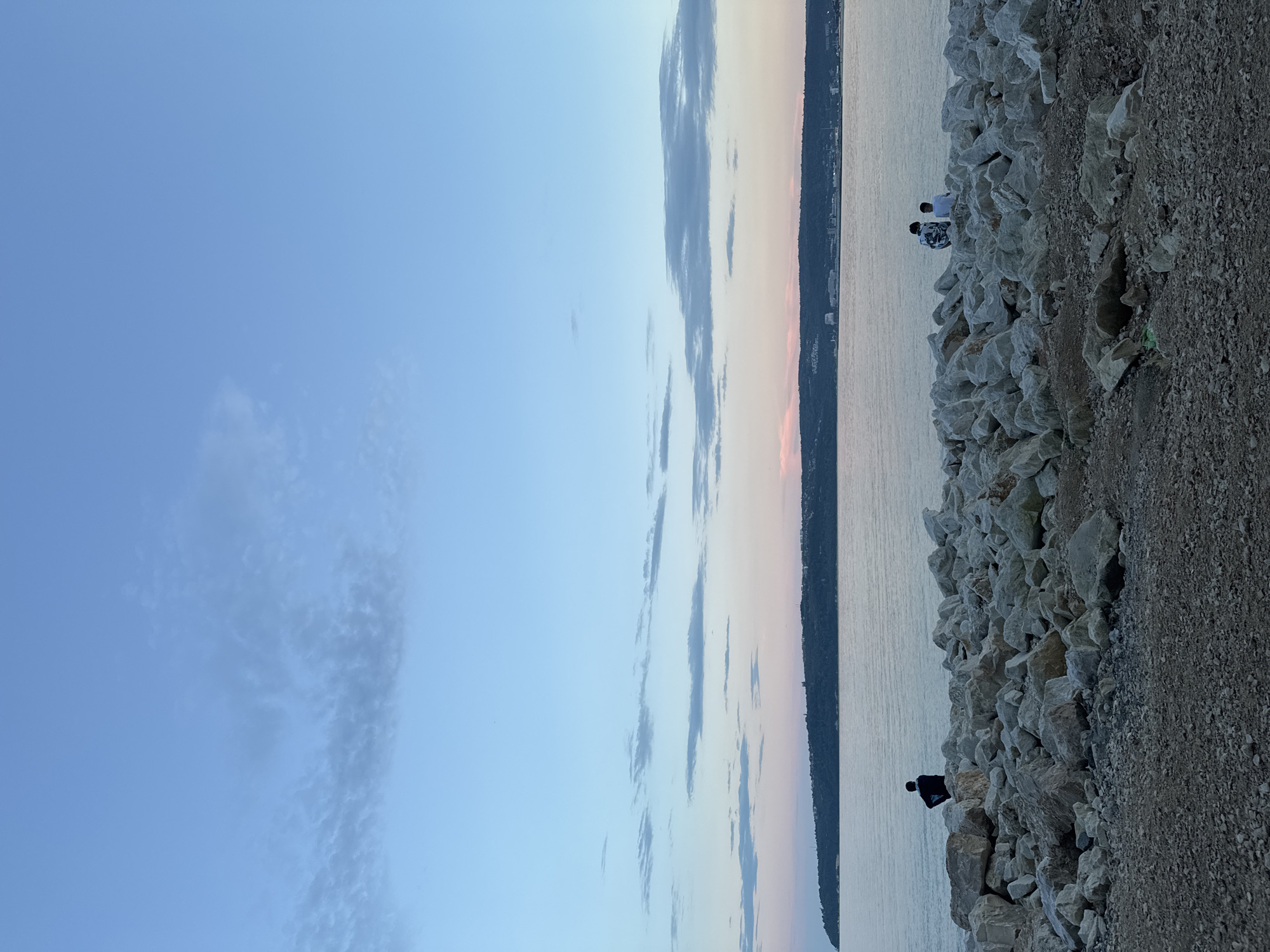 The image depicts a twilight coastal scene in Unknown, Unknown. The sky transitions from a clear, light blue at the top to a pale pink and orange hue at the horizon, with scattered, thin stratocumulus clouds stretching horizontally across the mid-sky. A distant, undulating landmass, possibly a coastline or island, is visible across the horizon, appearing dark green to bluish, with sparsely distributed structures or buildings along its crest and base. A calm body of water, reflecting the muted sky colors, occupies the mid-ground. The immediate foreground consists of a dark, gravelly ground surface, leading to a substantial formation of numerous irregularly shaped, light grey and white rocks, forming a natural or artificial breakwater. Three individuals are present on these rocks, facing the water with their backs to the viewer. One person is positioned on the left side of the frame, clad in dark attire. Two other individuals are seated together on the right side; one wears a patterned dark and white top, and the other wears a white top. The overall lighting suggests either sunrise or sunset.